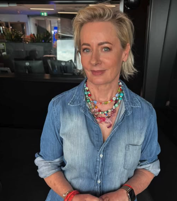 A woman with short blonde hair and light skin wearing a denim shirt, colorful necklaces, and bracelets, standing indoors.