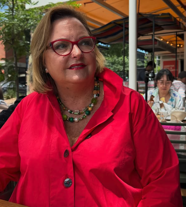 A woman with shoulder-length brown hair, wearing red glasses, a red blouse, and a multicolored beaded necklace, sitting outdoors at a restaurant or cafe with other diners in the background.
