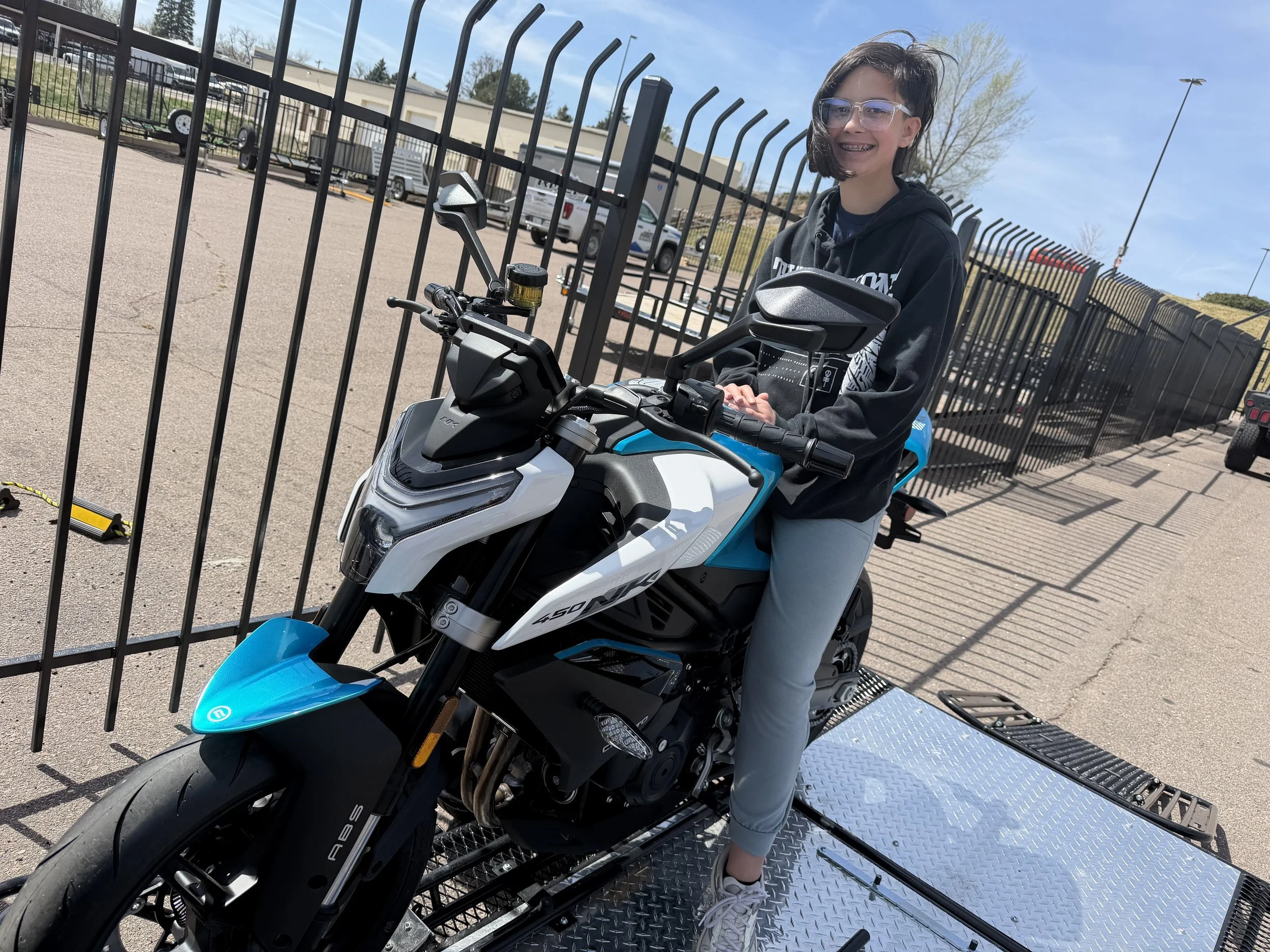 A young woman with glasses and short hair sitting on a white and black electric motorcycle, smiling outdoors near a metal fence with parked vehicles in the background.