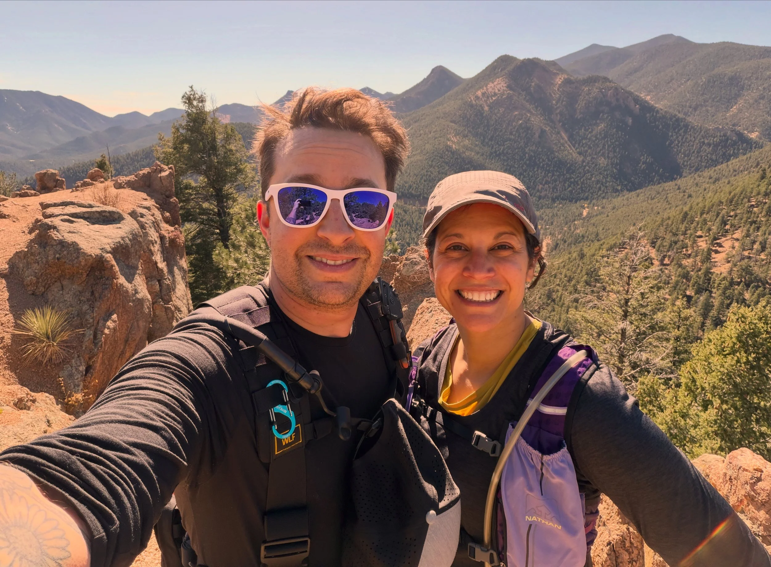 A smiling man and woman taking a selfie on a mountain trail with a background of mountain ranges, trees, and rocky terrain on a sunny day.