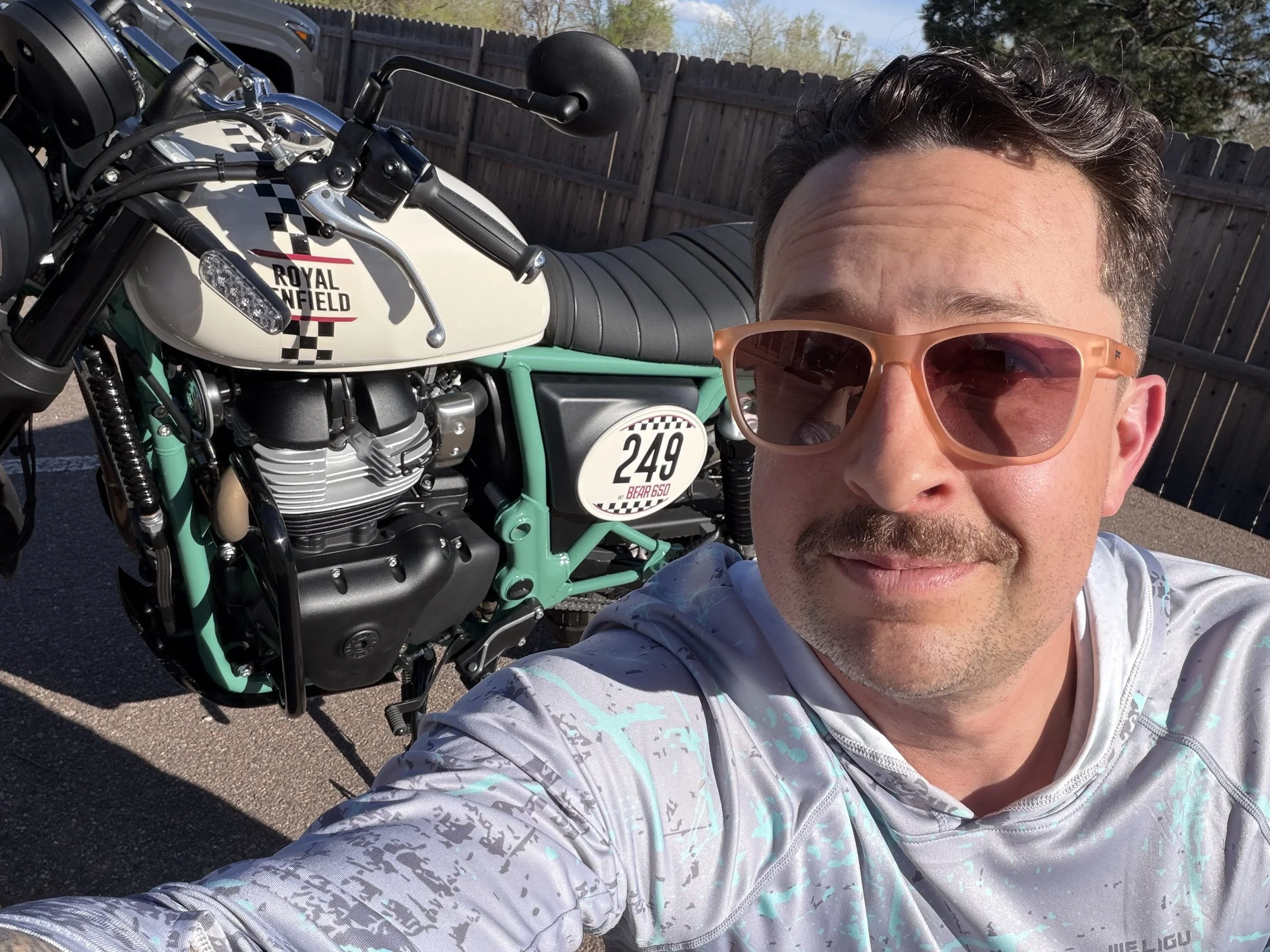 Man wearing pink sunglasses takes a selfie in front of a vintage style Royal Enfield motorcycle with a racing number 249, parked on a paved area with a wooden fence and trees in the background.