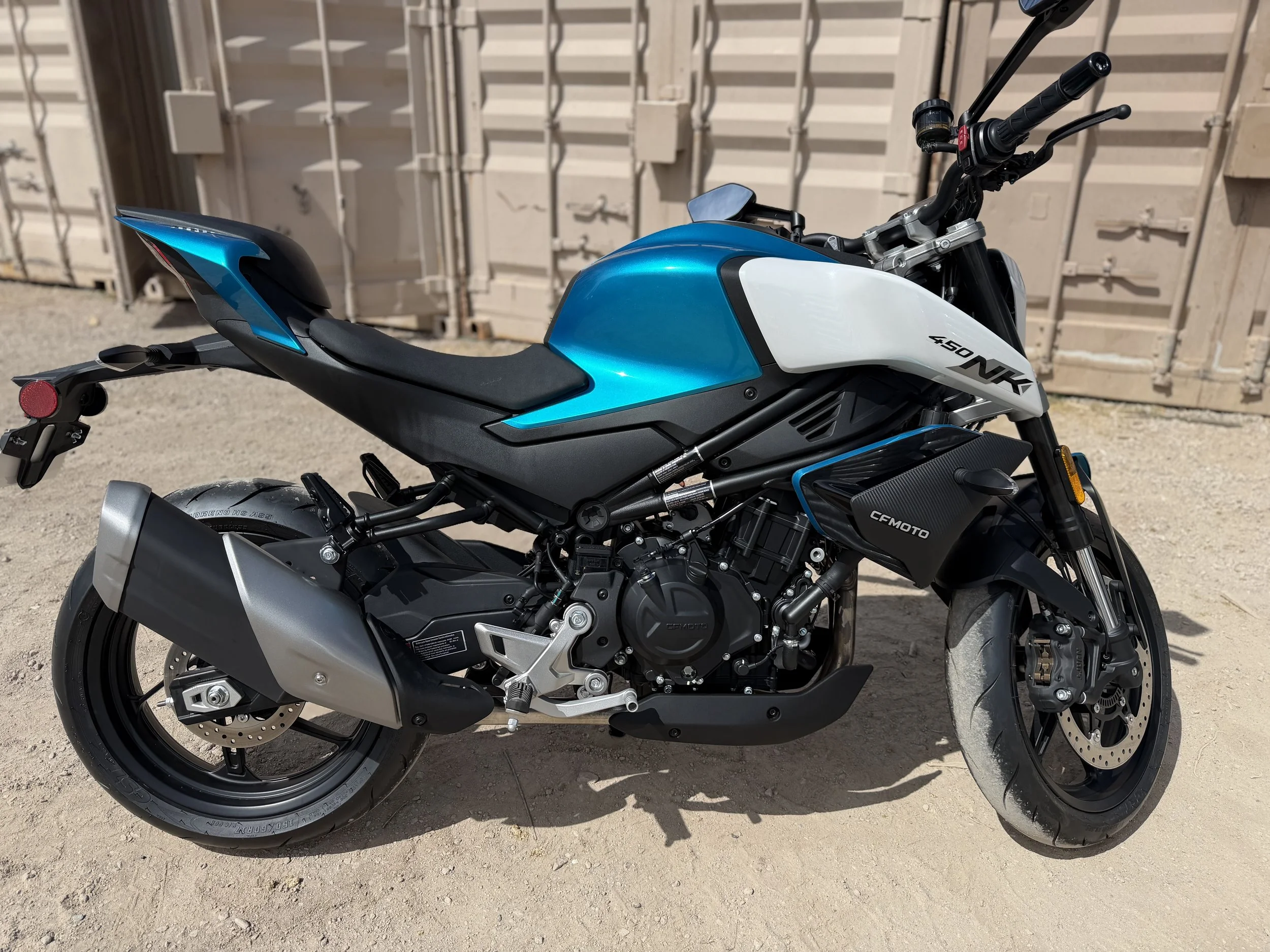 Blue and white electric motorcycle with black accents parked on a dirt surface in front of a beige industrial container.