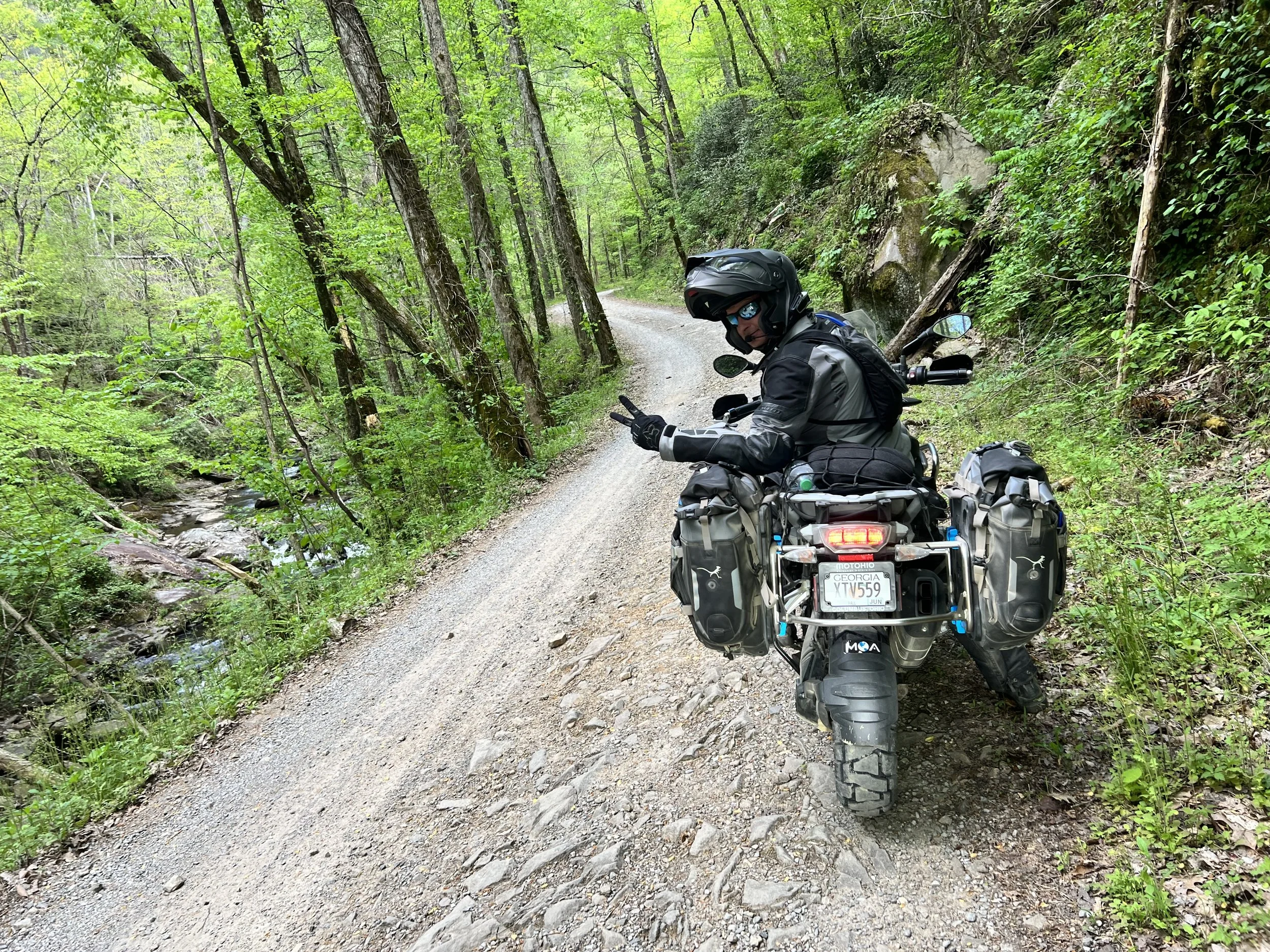 A motorcyclist wearing a helmet, sunglasses, and motorcycle gear sitting on a touring bike on a dirt trail surrounded by green trees and a small creek to the left.