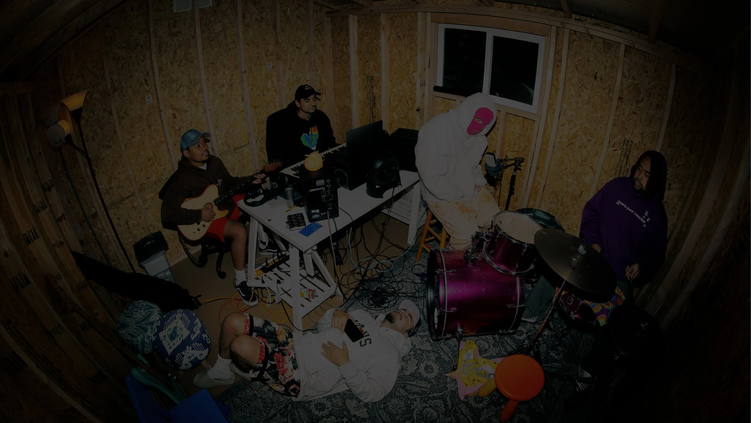 A group of five people in a music studio room with wooden walls. One person is lying on the floor holding a phone, another is playing an electric guitar, a third is sitting at a keyboard, a fourth is sitting behind a drum set, and the fifth is standing in front of the drums. The room has various musical equipment and toys.