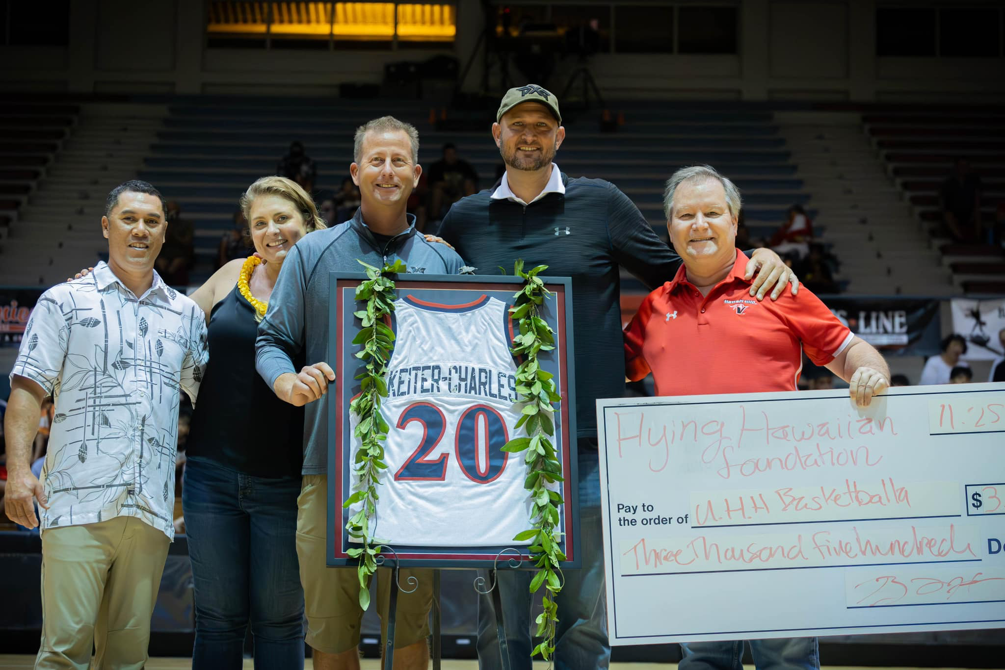 Group of five people standing on a basketball court celebrating a donation, holding a framed jersey with number 20 and a giant check for $3,000 made out to U.H.H. Basketball, decorated with greenery.