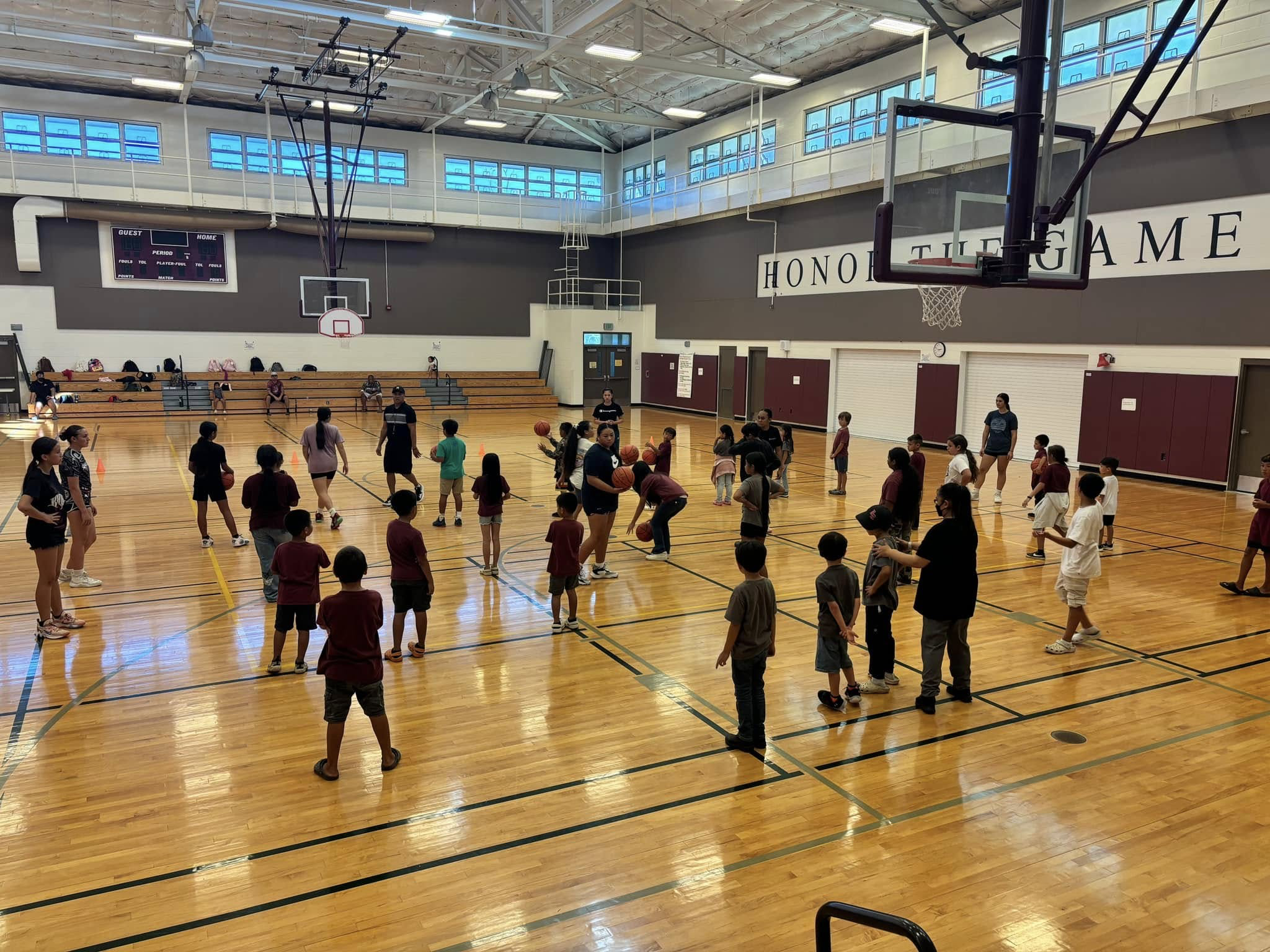 Children participating in a basketball class in a gymnasium with coaches, with some sitting on bleachers and a large scoreboard.