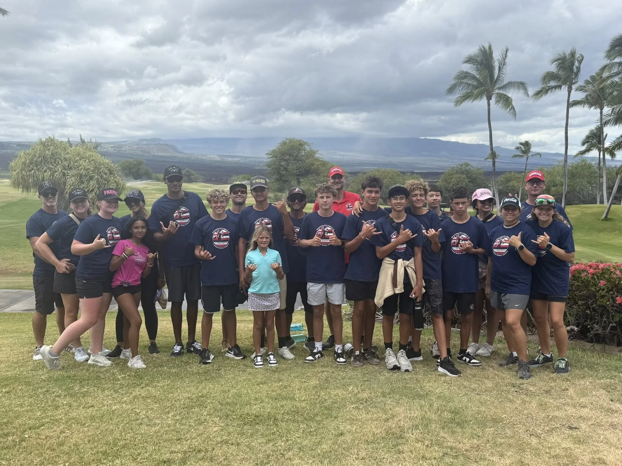 A group of children and young adults posing on a golf course under cloudy skies, with palm trees and mountains in the background, some making hand gestures.