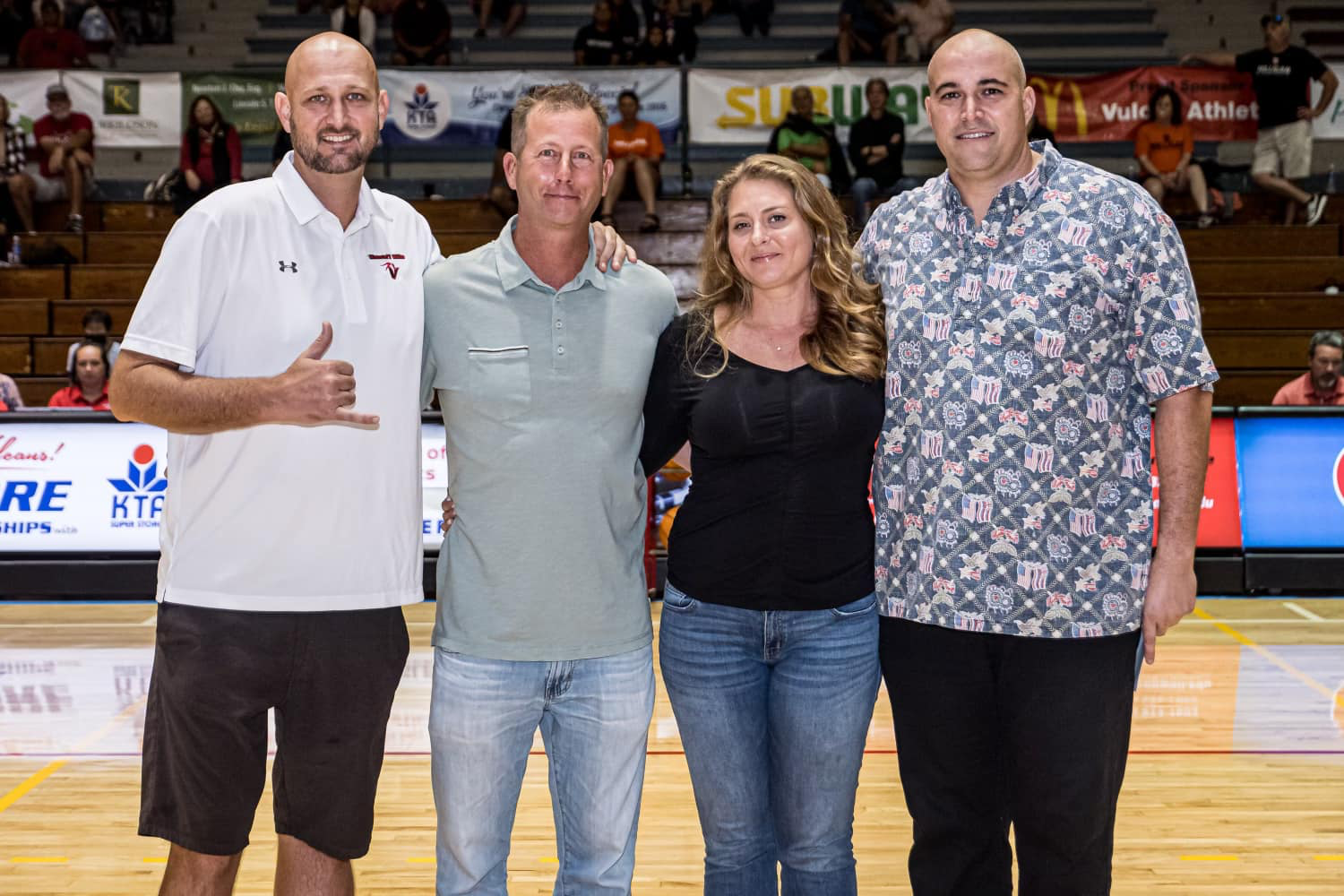 Four people standing on a basketball court, posing for a photo, with audience in the background.