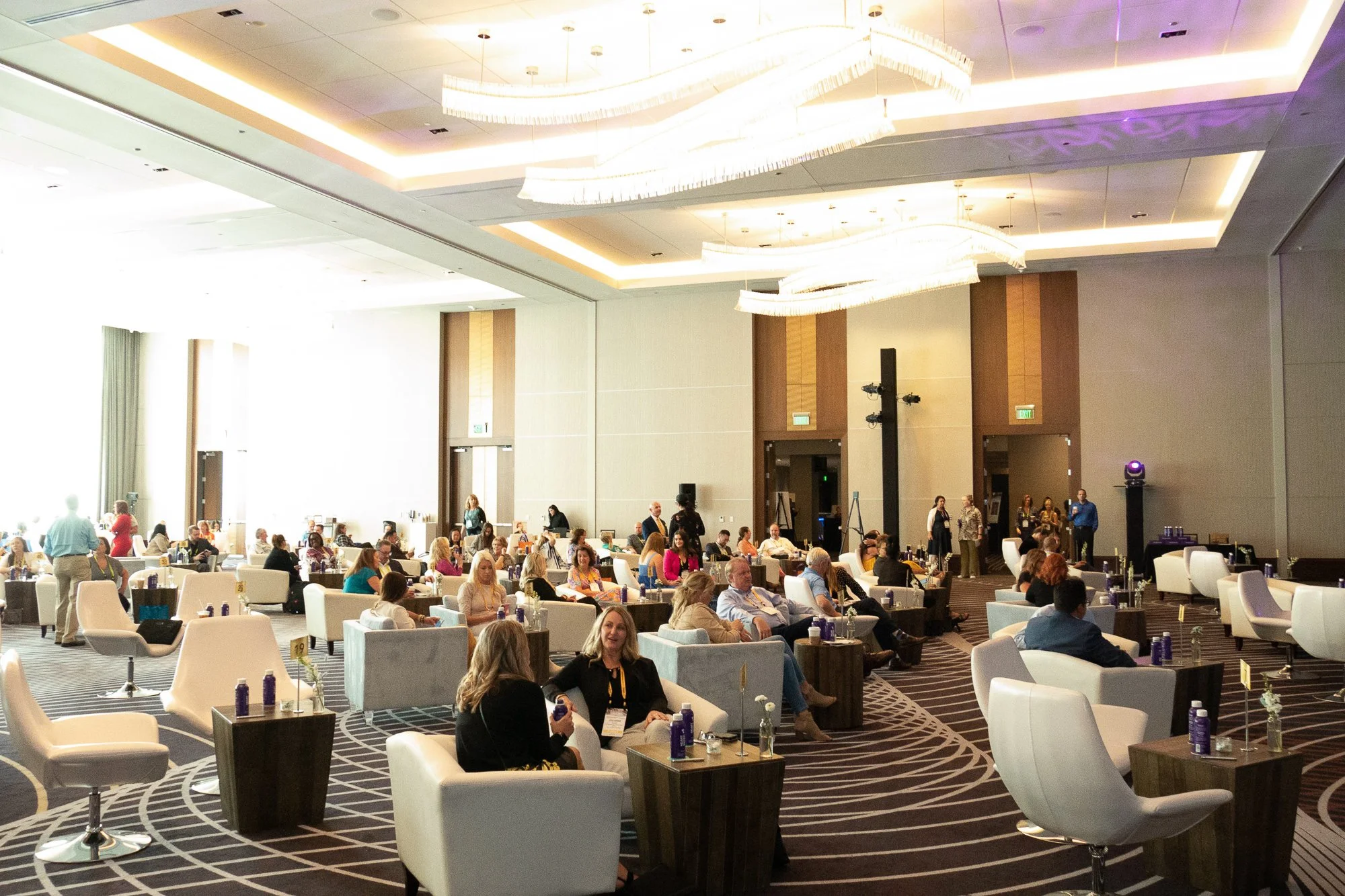 A large, well-lit conference room filled with people seated in white armchairs and standing, with tables on some. The ceiling has elegant, modern chandeliers, and the room features a patterned carpet and a high ceiling with light fixtures. Attendees appear engaged, with some wearing badges, and there are water bottles on tables.