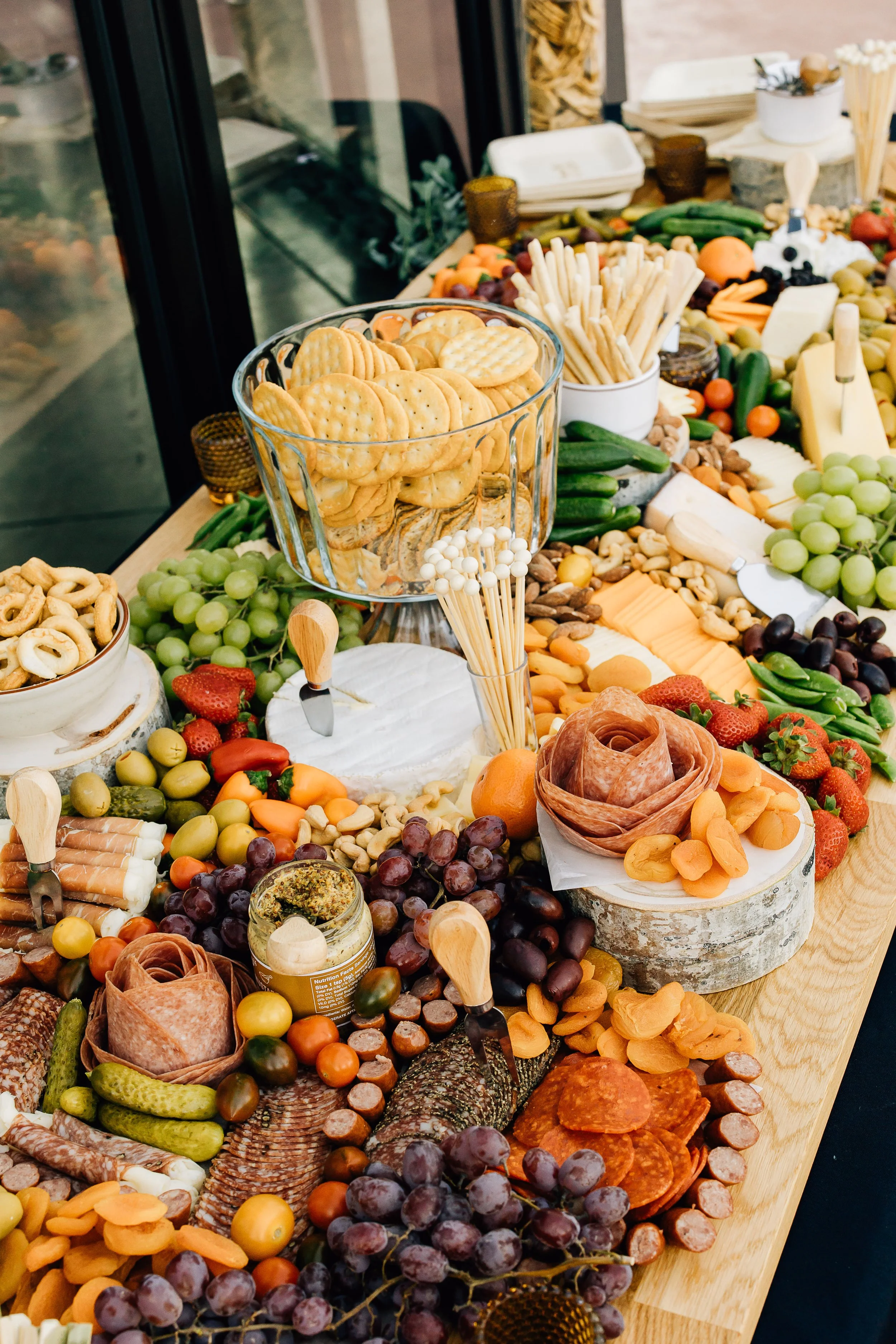 A spread of assorted snacks including crackers, cured meats, cheeses, grapes, cherry tomatoes, and various vegetables on a wooden table.