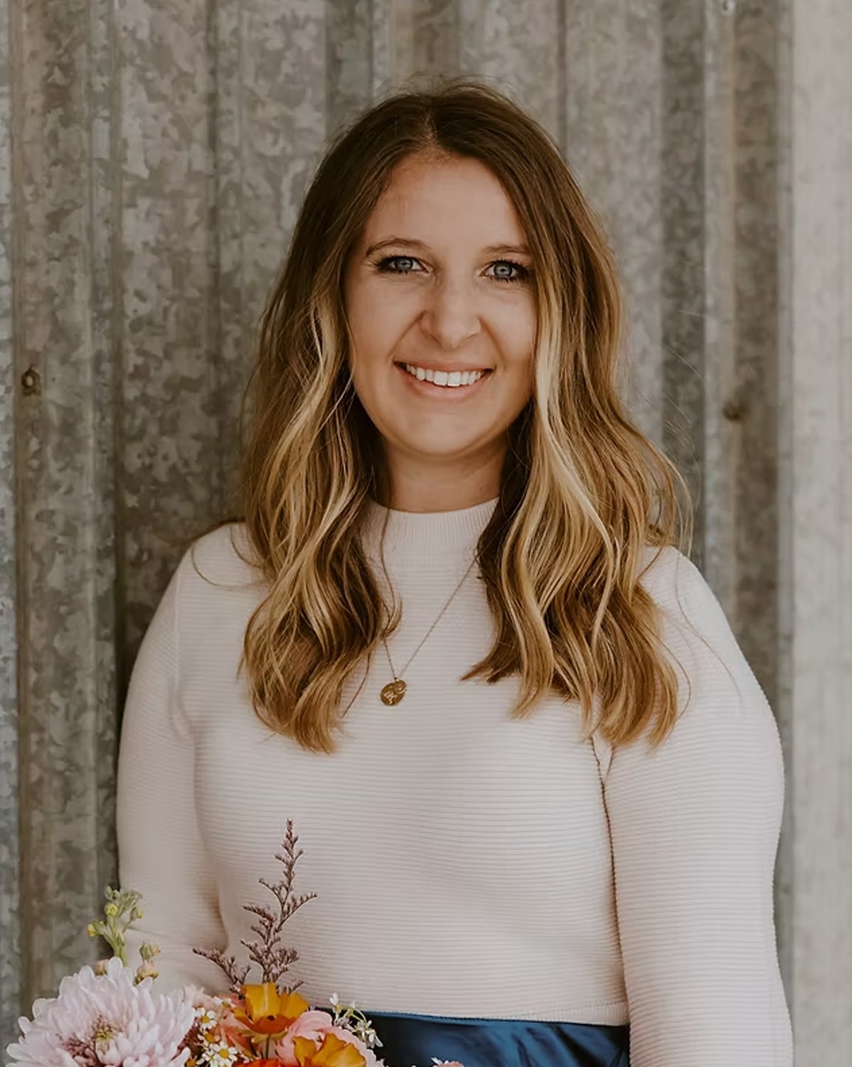 A woman with wavy, light brown hair and blue eyes smiling in front of a rustic wooden background, wearing a cream-colored long-sleeve top, a gold necklace, and holding a colorful bouquet of flowers.