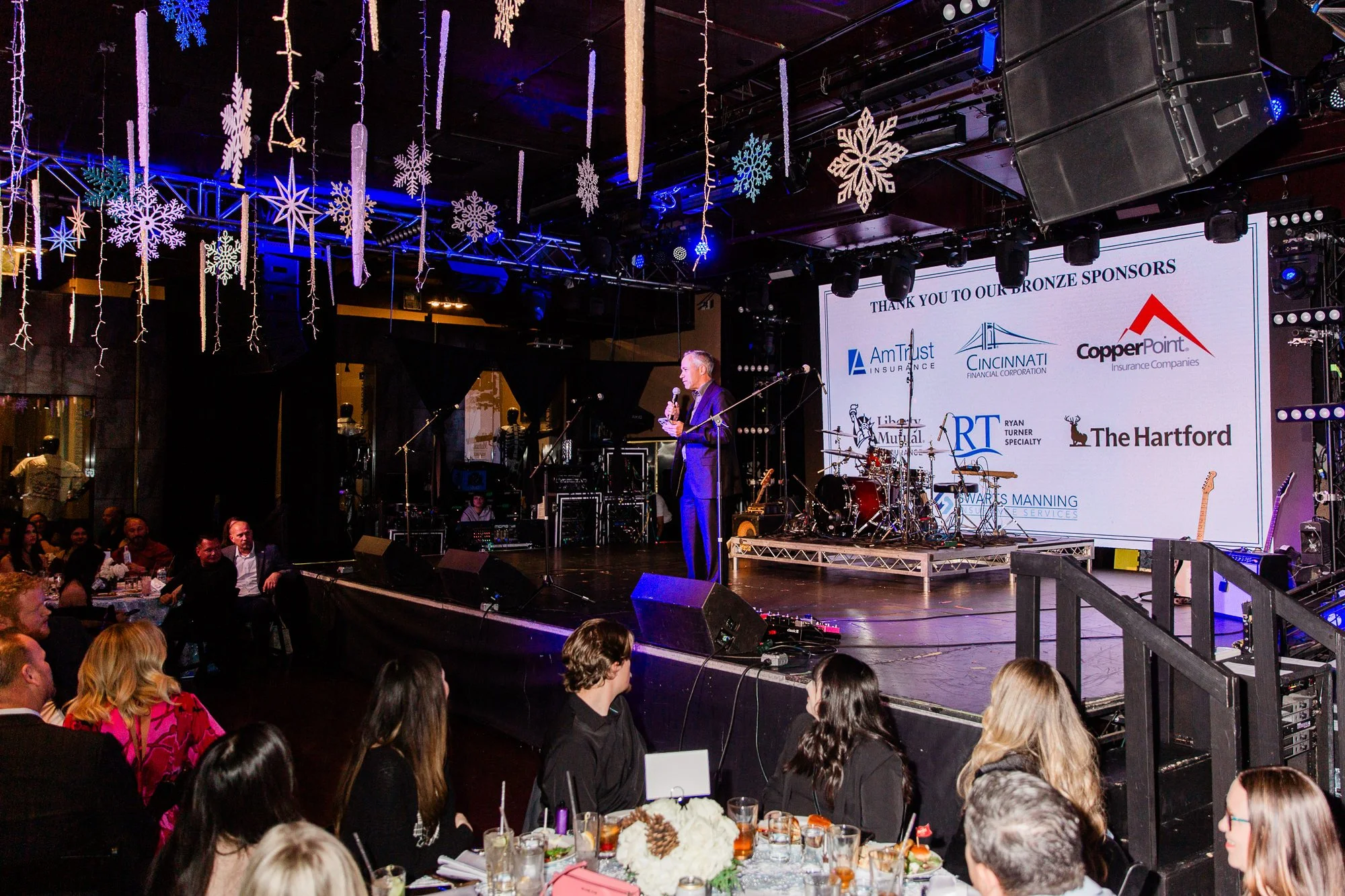 A stage with a speaker, musical instruments, and a large screen displaying sponsor logos at a formal event. There are holiday decorations hanging from the ceiling, including snowflakes and icicles. The audience, seated at tables with food and drinks, watches the event.