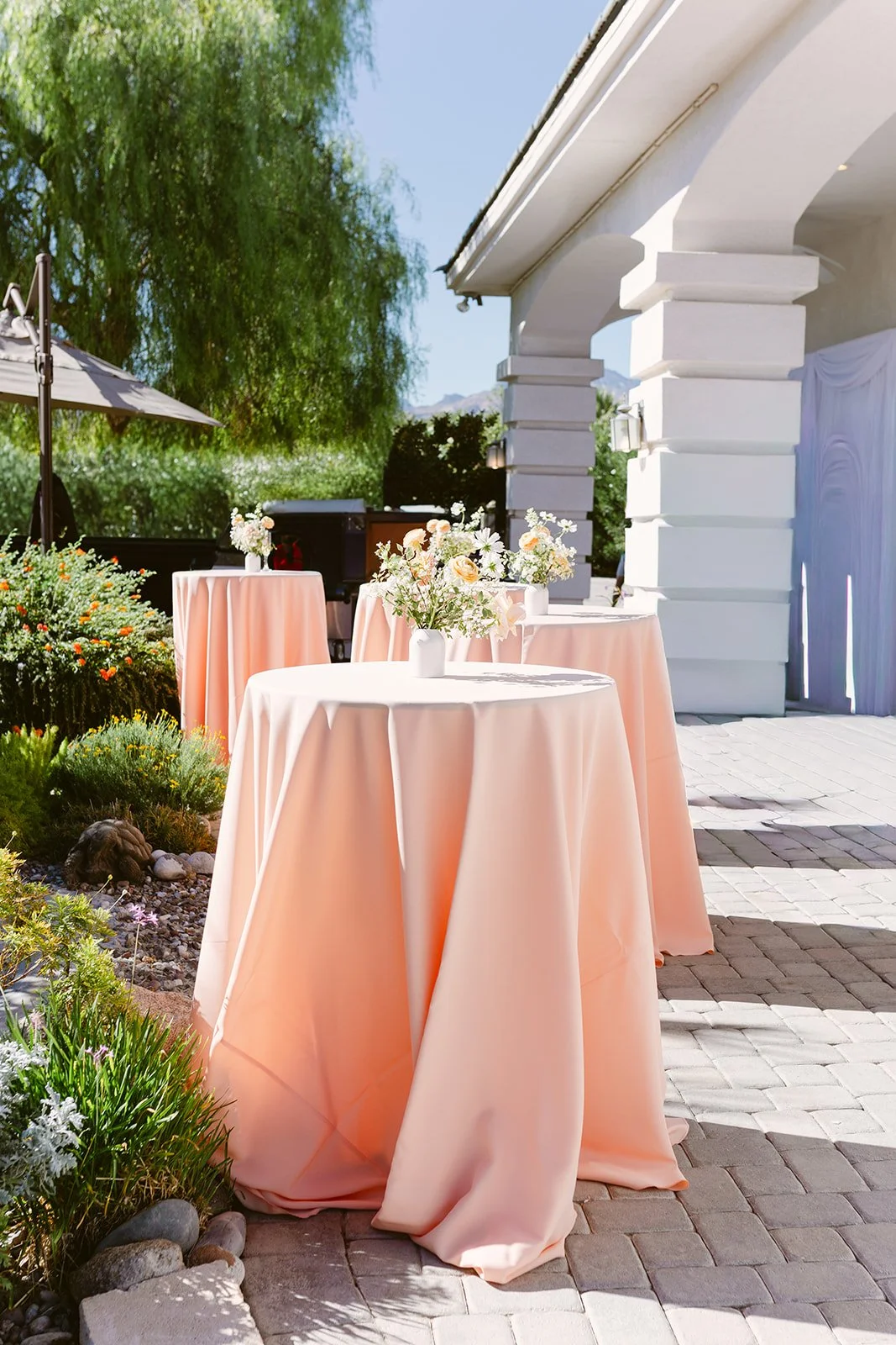 Outdoor event space with round tables covered in pink tablecloths and decorated with floral centerpieces, adjacent to a white building with large columns, surrounded by greenery and orange flowers under a partly cloudy sky.