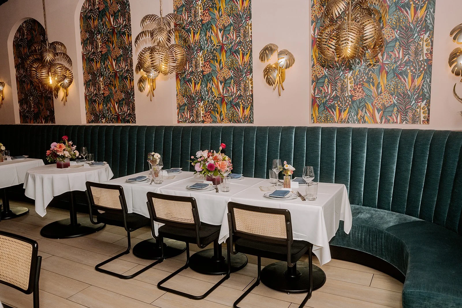 An elegant restaurant dining area with a long U-shaped dark green velvet banquette, a white tablecloth-covered table set with glassware, plates, napkins, and floral centerpieces, and wall decor featuring colorful tropical-patterned wallpaper and gold leaf-shaped wall sconces.