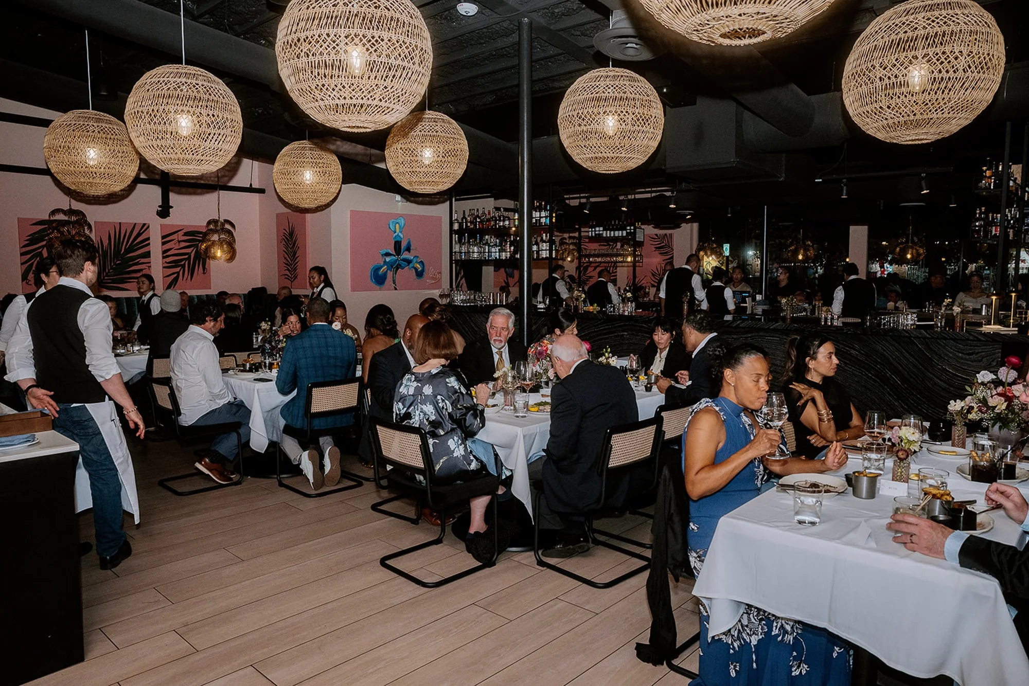 A crowded restaurant with patrons seated at tables, waiters serving food, and a bar area in the background. The decor features large, woven, round hanging light fixtures and pink wall art with botanical designs.