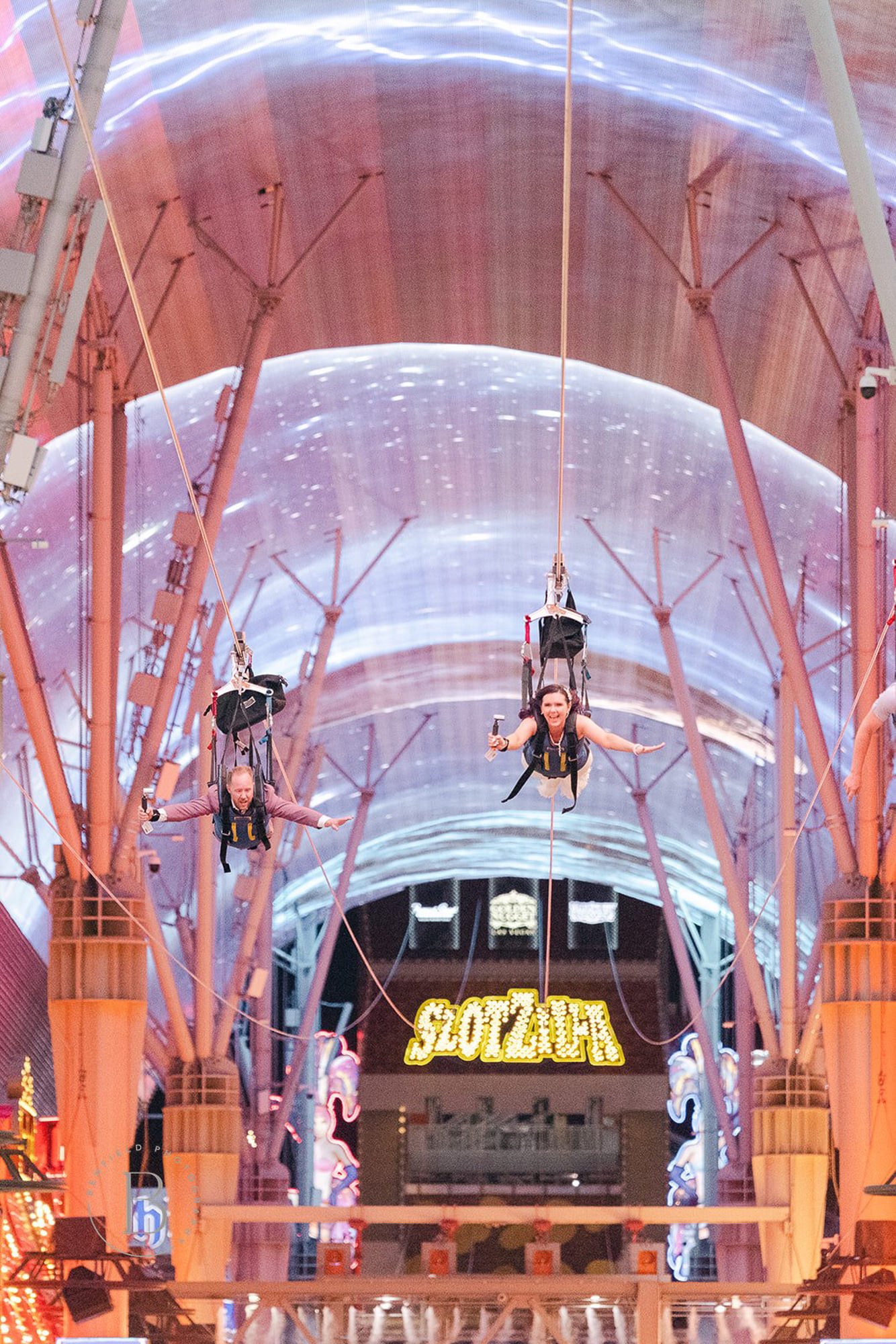 People riding zip lines inside a colorful, illuminated arcade or entertainment area. The ceiling features a neon sign that reads 'SLOT & LAND' and decorative dragon figures.