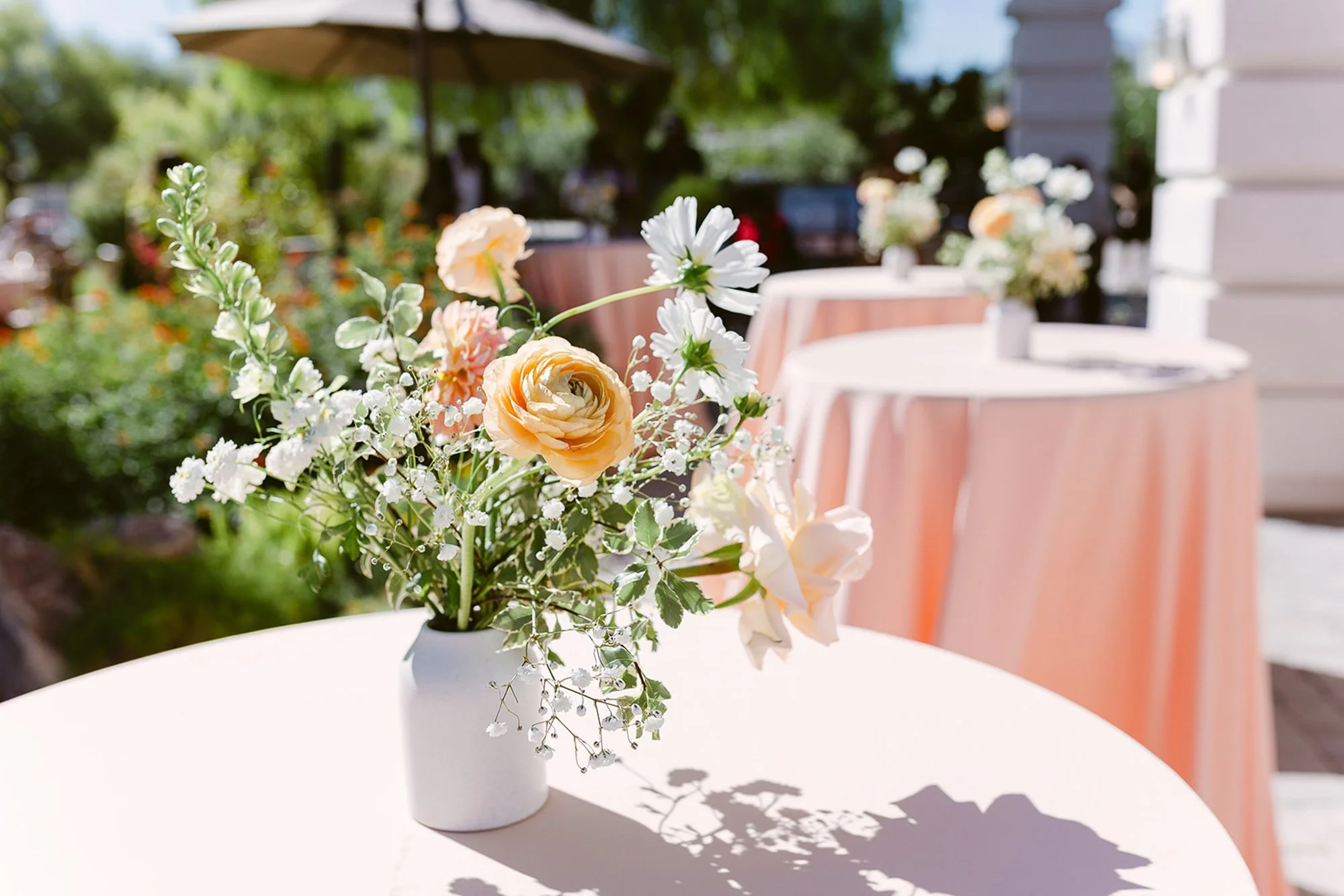 A white vase containing a bouquet of peach and white flowers on a white table outdoors on a sunny day.