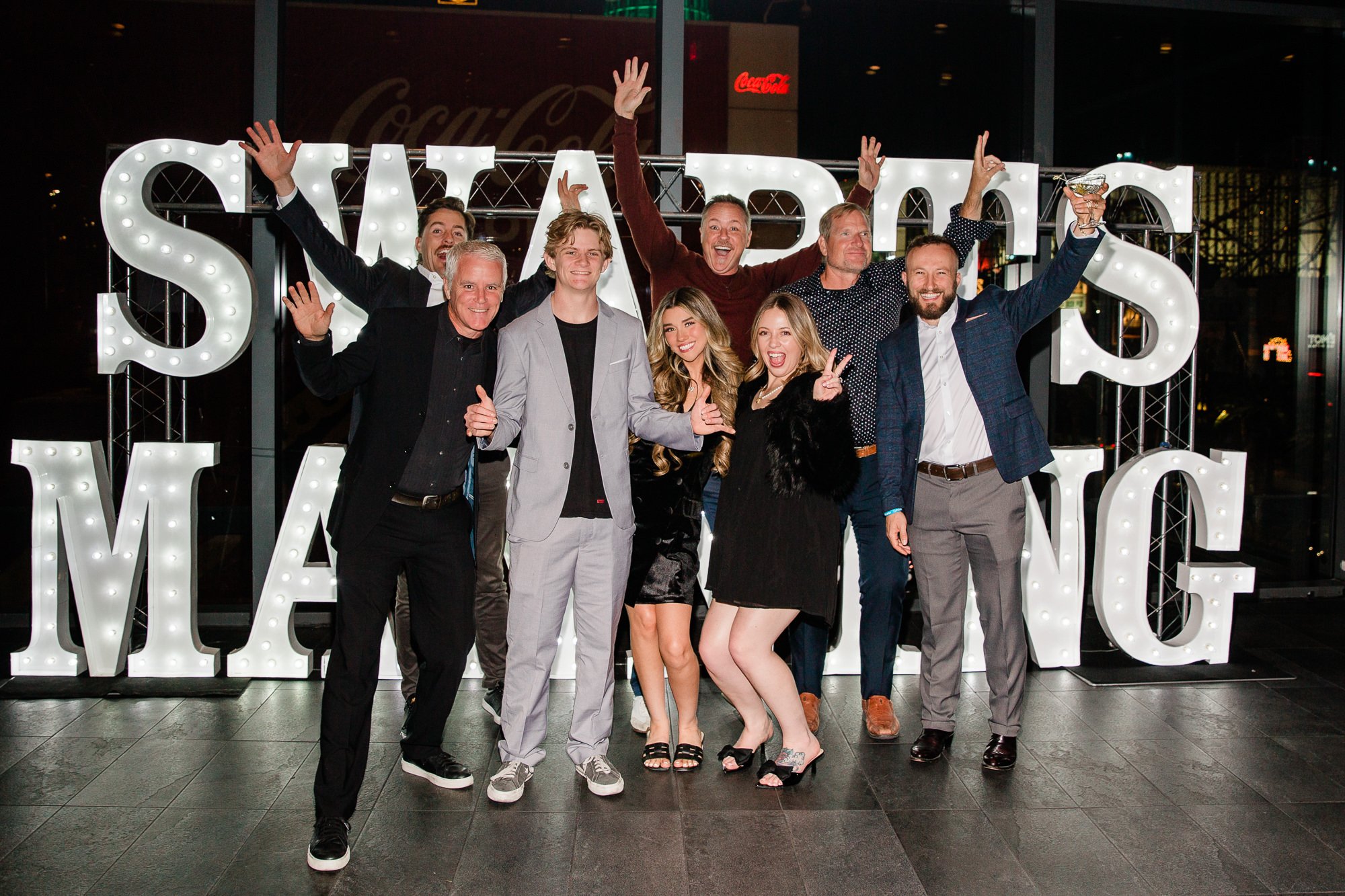 A group of nine people celebrating at night, standing in front of a large illuminated sign with the words 'Sorry' and 'W' visible. They are smiling, with some raising their hands and holding drinks, dressed in formal and semi-formal attire, inside a modern building.
