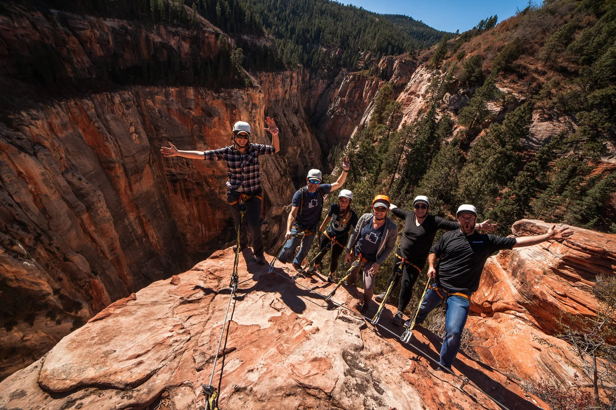 Group of seven people standing on a rock ledge with a canyon and forest in the background, all wearing helmets and climbing gear, smiling and posing for a photo during an outdoor adventure.