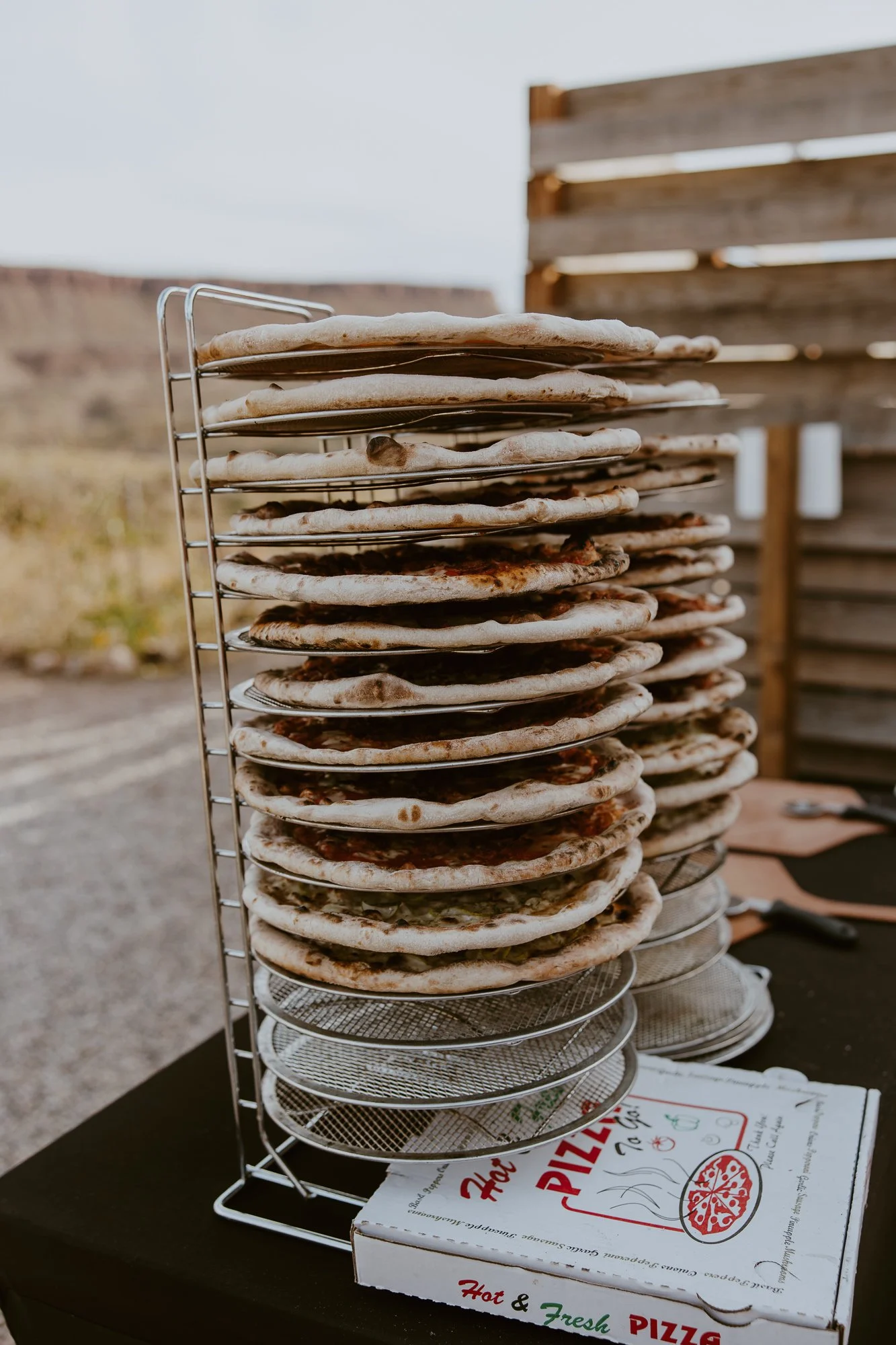 Stack of cooked pizzas cooling on a metal rack outdoors, with a pizza box on a table nearby and a rustic wooden fence in the background.
