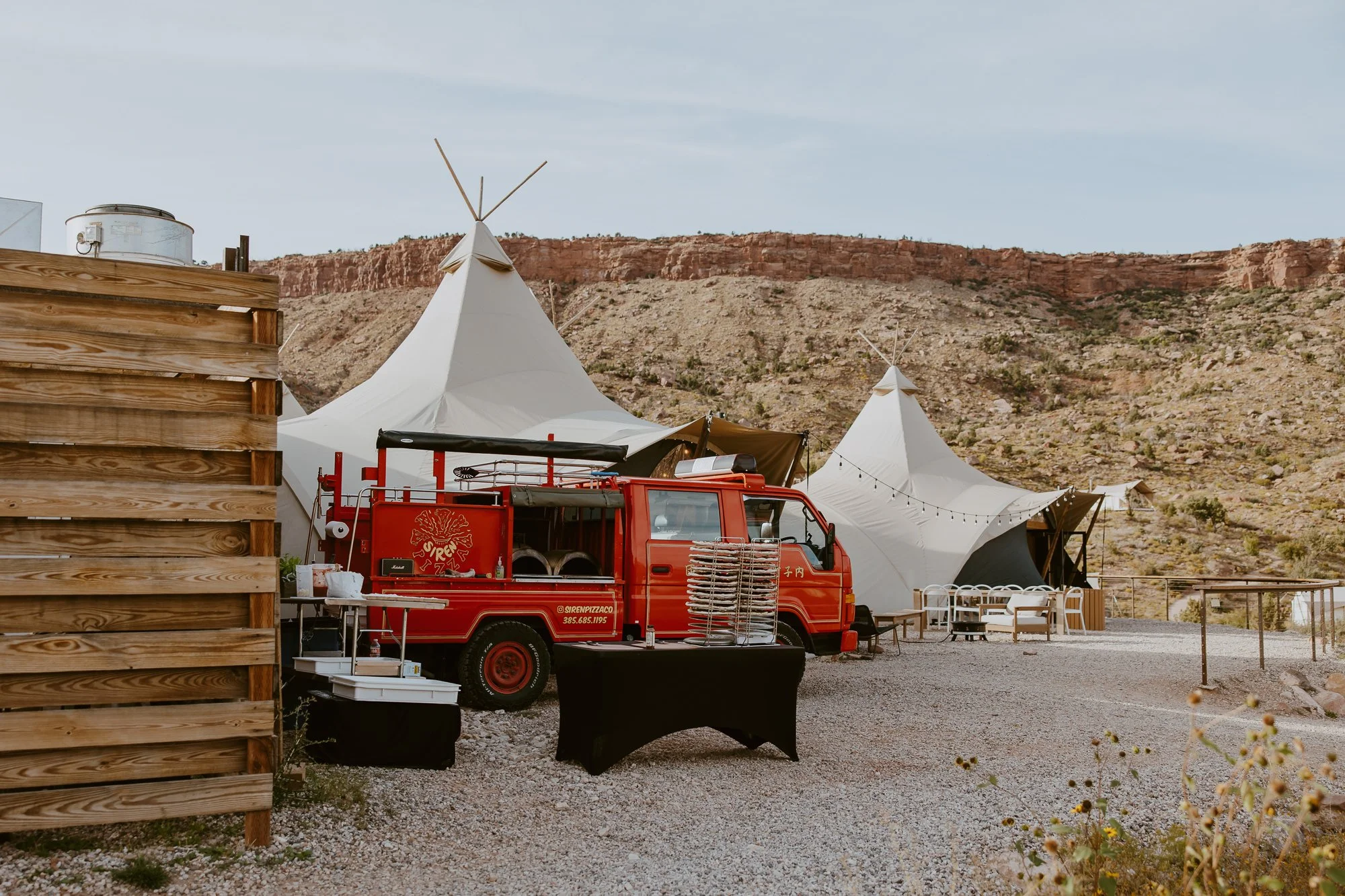 Outdoor scene with white tents, a red food truck, and a wooden fence in a desert landscape with rocky hills in the background.