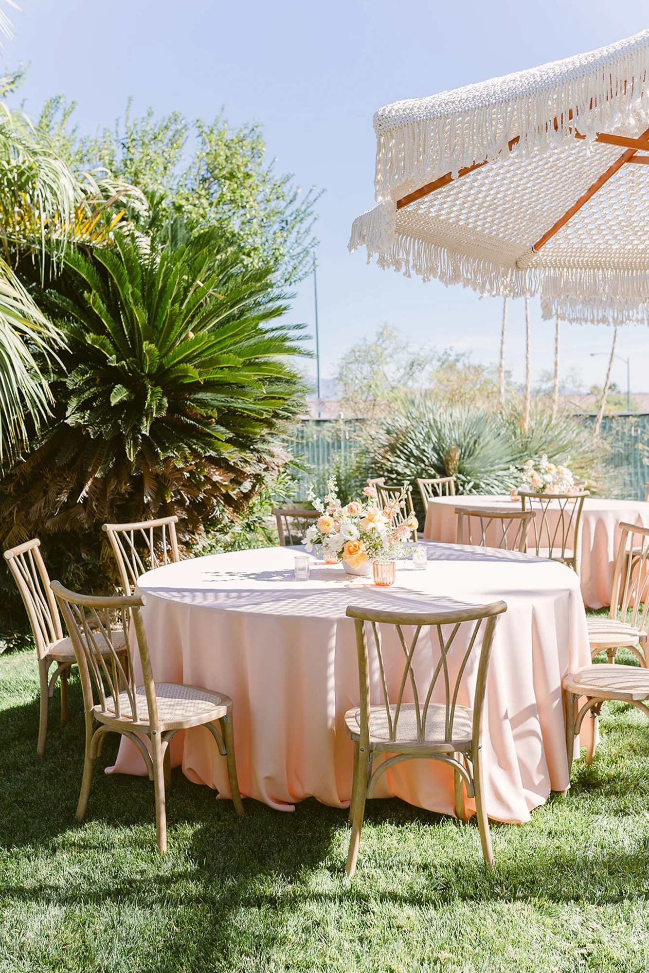 Outdoor setup with a round table covered in a pink tablecloth, surrounded by wooden chairs with light-colored cushions, floral centerpiece, under a white layered canopy, in a grassy area with lush green trees and plants, sunny sky.