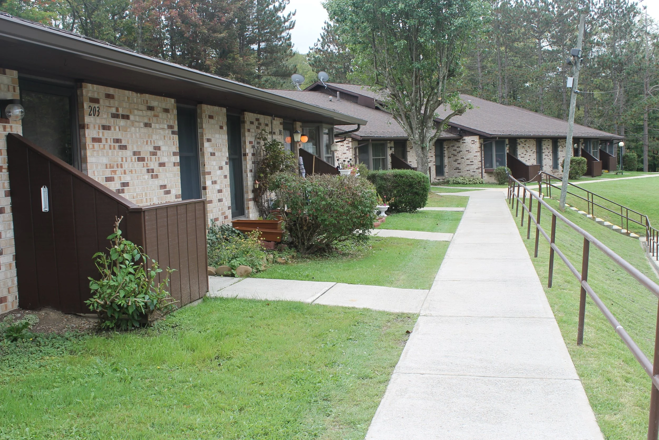 Pineview Apartment complex with brick buildings, a sidewalk, trimmed grass, bushes, and trees, with a gentle slope and a railing along an incline.