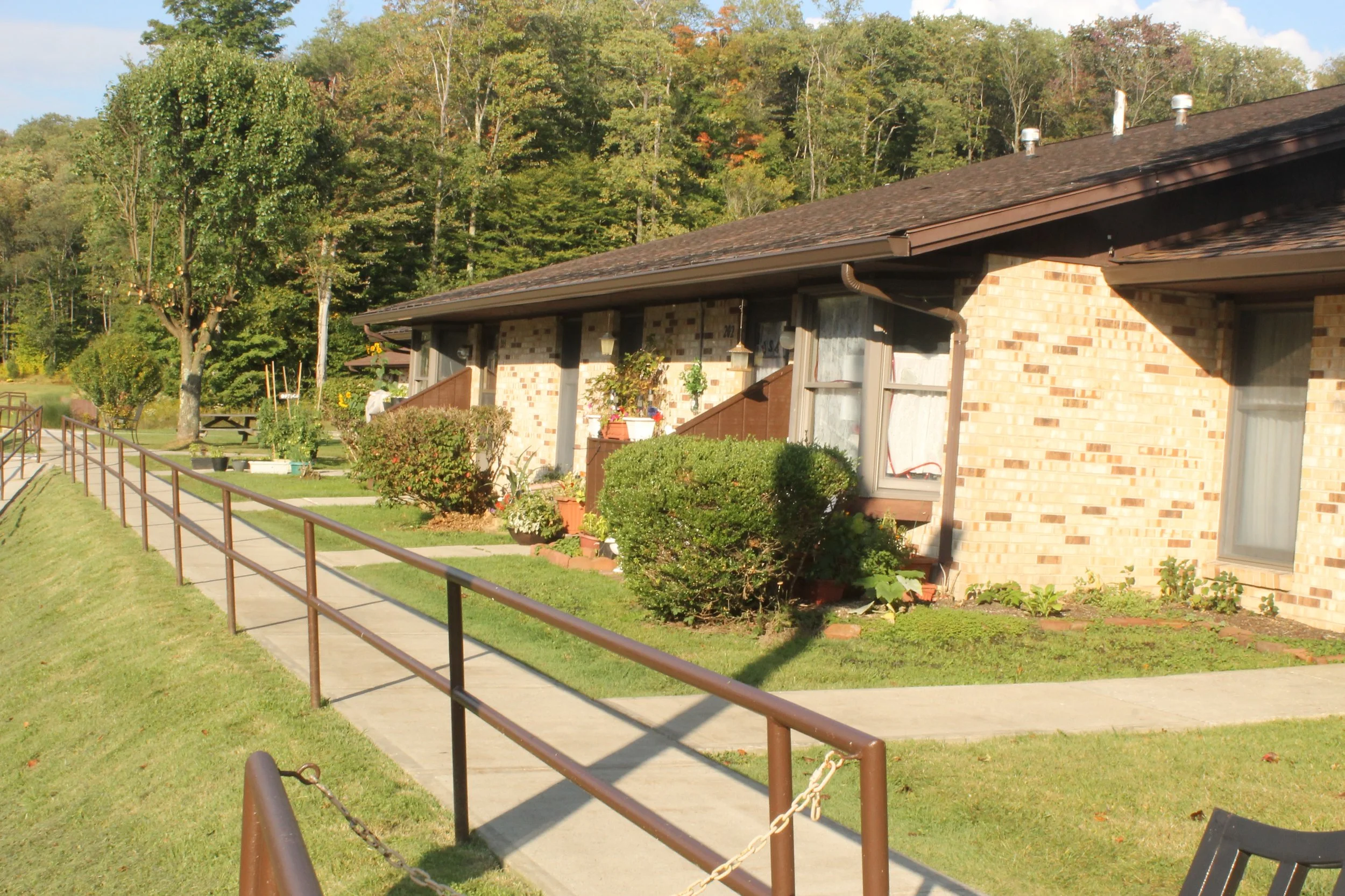 A row of single-story brick apartments at Pineview Apartments with small front porches and landscaped bushes. A concrete walkway runs parallel to the building, with a metal handrail along the sidewalk. 