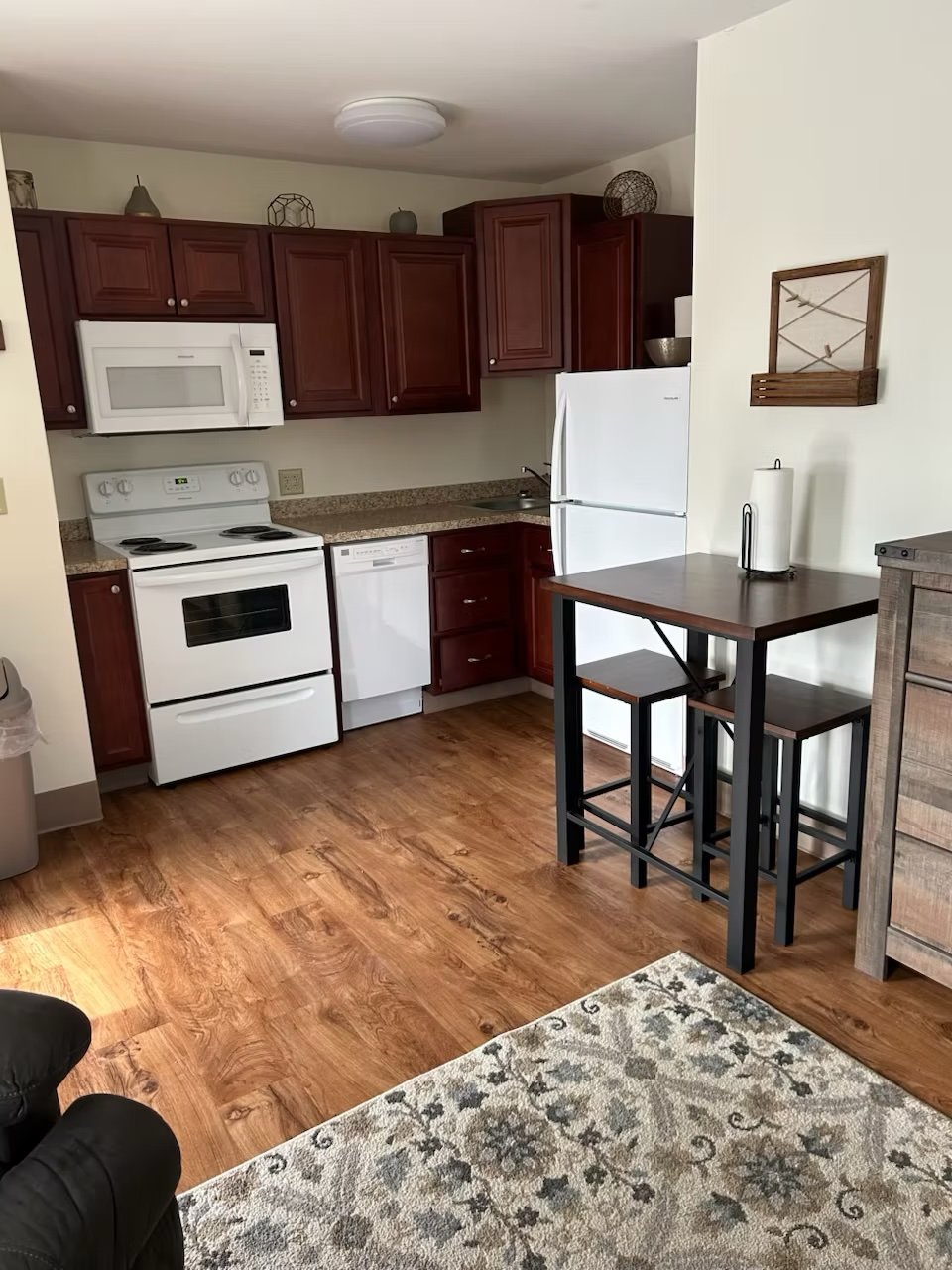 A Rapids Apartment studio with kitchen containing white appliances, cherry wood cabinets, wood flooring, small dining table with two chairs, and decorative items on top of cabinets.