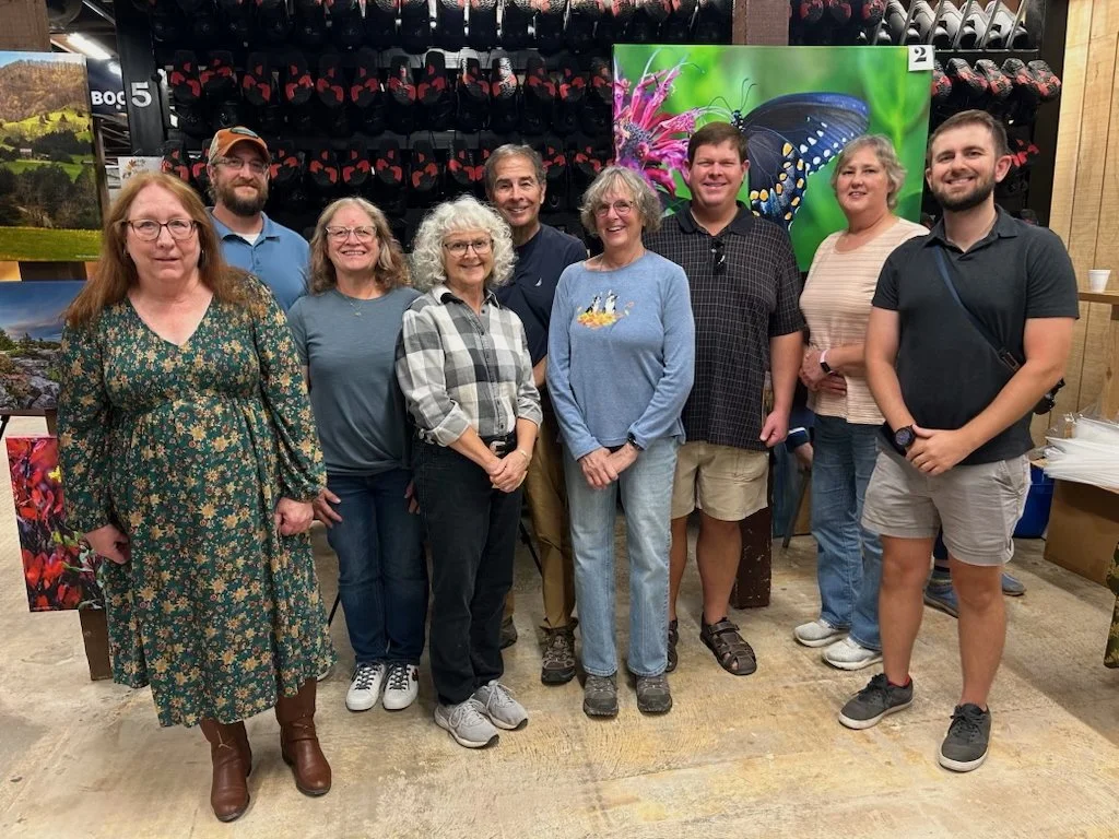 Group of winning photographers from Blackwater Ridge Foundation's photo contest standing together in front of a display of paintings and a butterfly photograph, in Timberline Ski Lodge during Artoberfest debut.