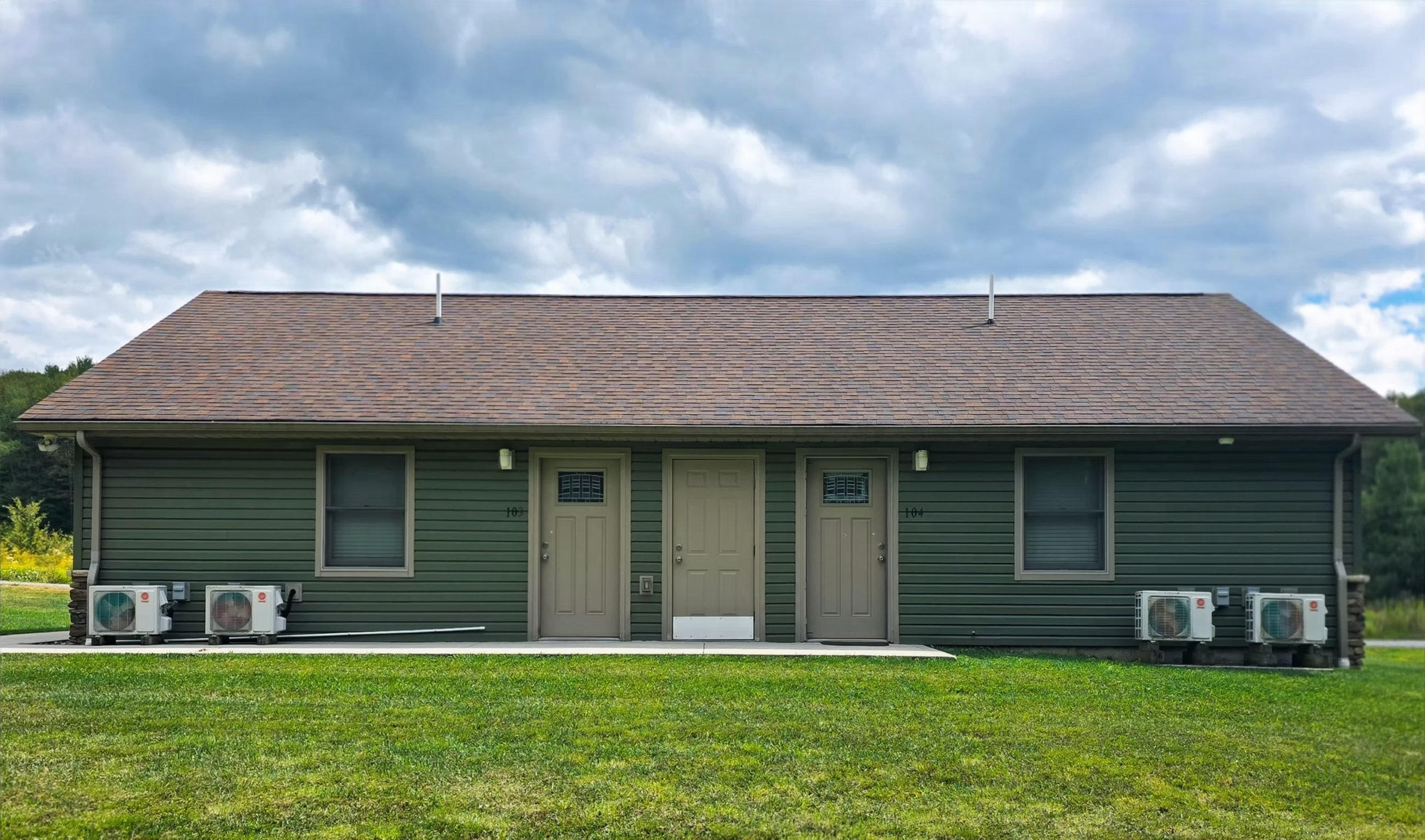 This is an example of our Rapids apartment building. Each is  single-story building with green siding, external front doors to each apartment, windows, and a grassy yard and cloudy sky. Flexible leases and fully furnished make each apartment perfect.