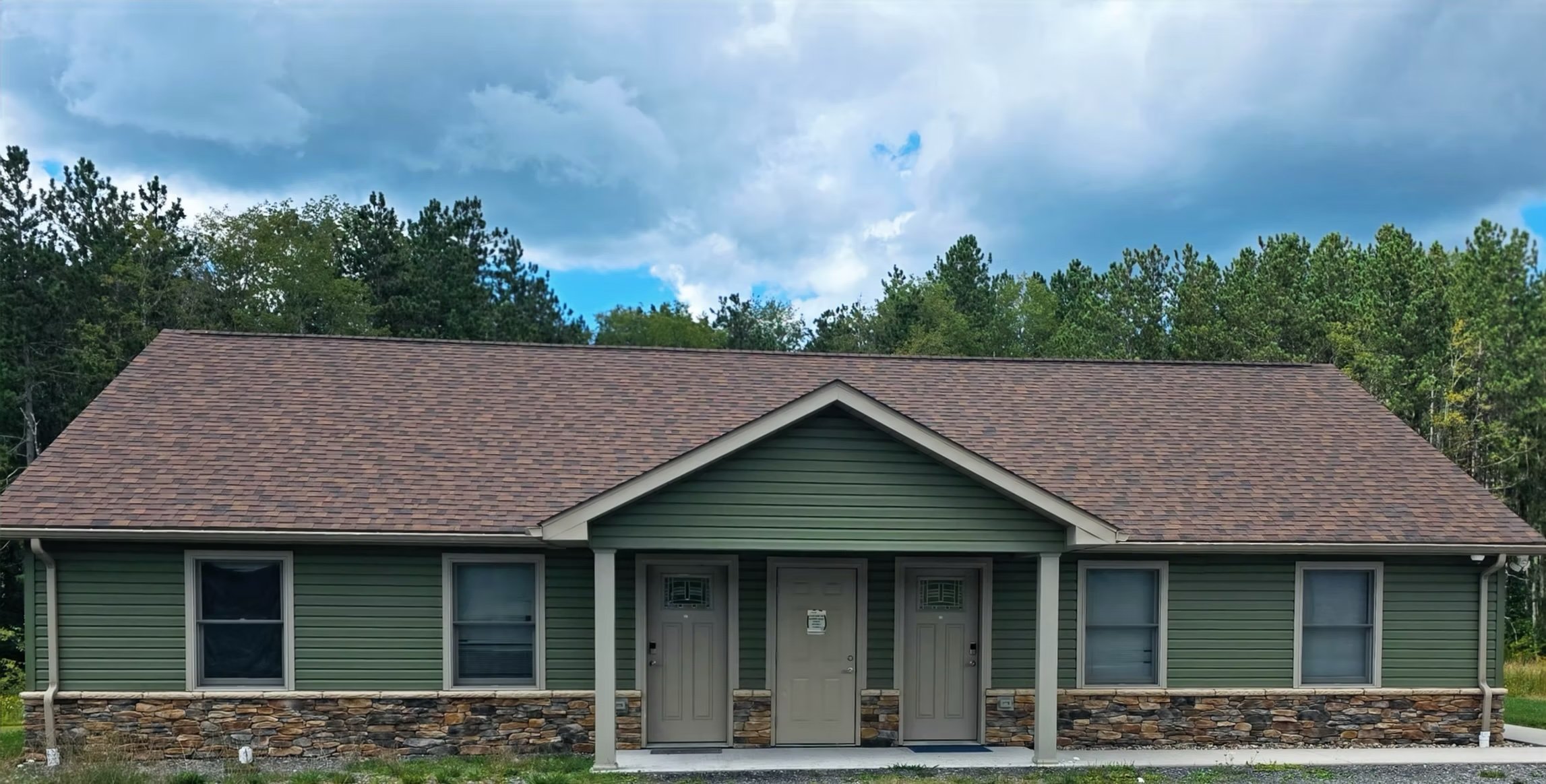 A single-story Rapids apartment building: green house with stone foundation, three front doors, and stone steps, surrounded by green trees under a partly cloudy sky.