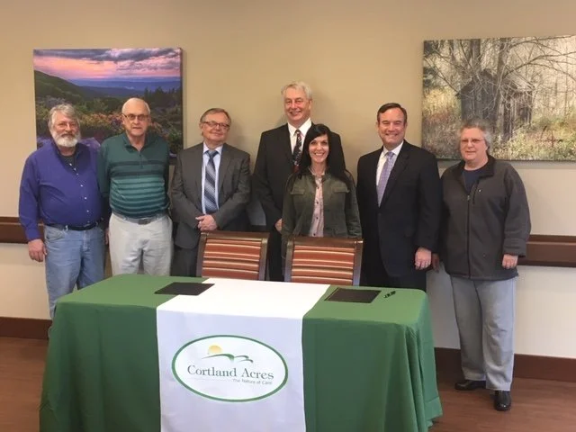 Group of board members and staff standing behind a table with a sign that reads Cortland Acres, in a room with landscape paintings on the walls. The  history of Cortland Acres runs back decades.