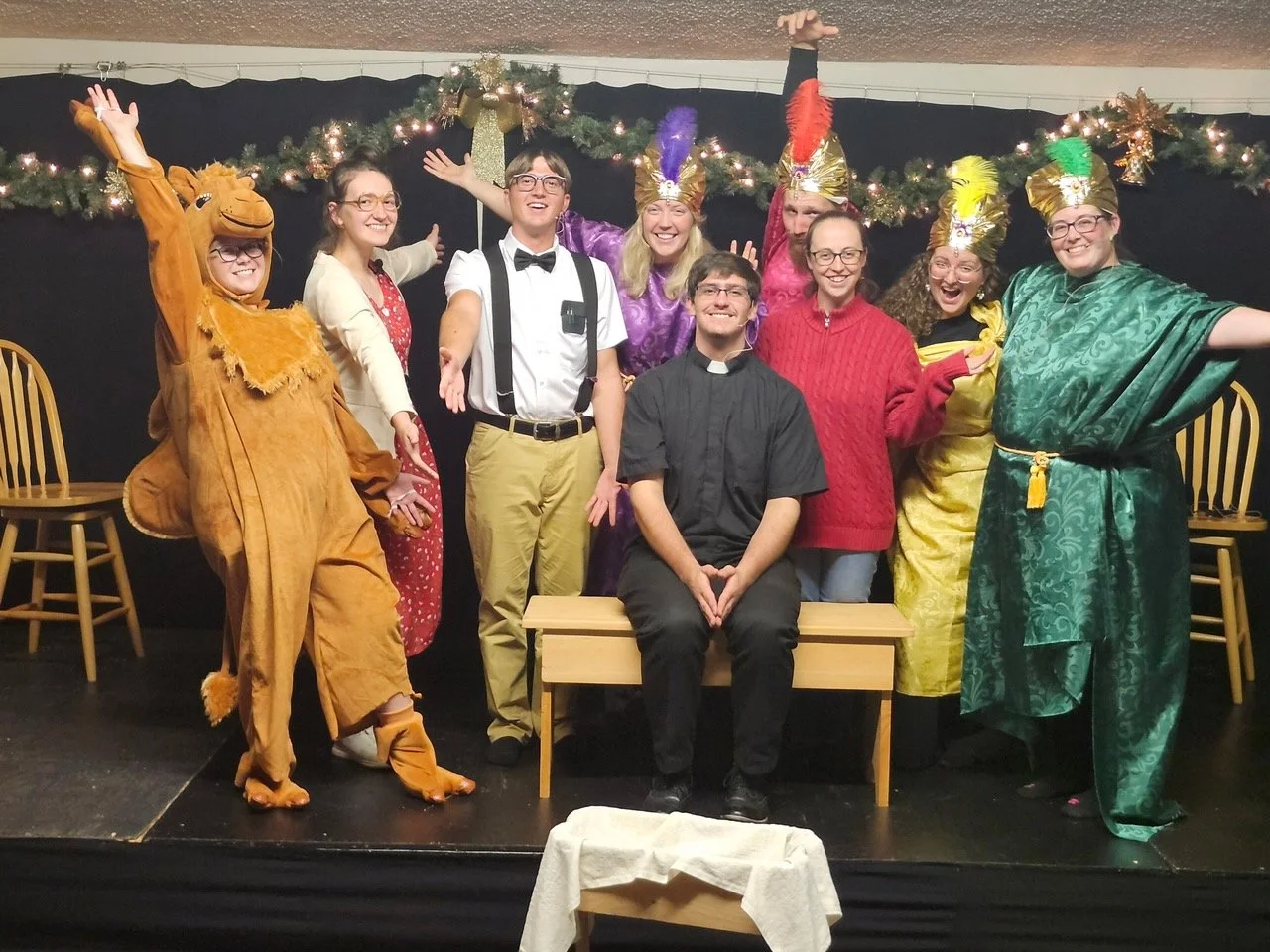 Group of actors from benefit dinner dressed in costumes, including a person in a lion costume, others in colorful and festive attire, standing on a stage with Christmas decorations in the background. One person is seated on a bench.