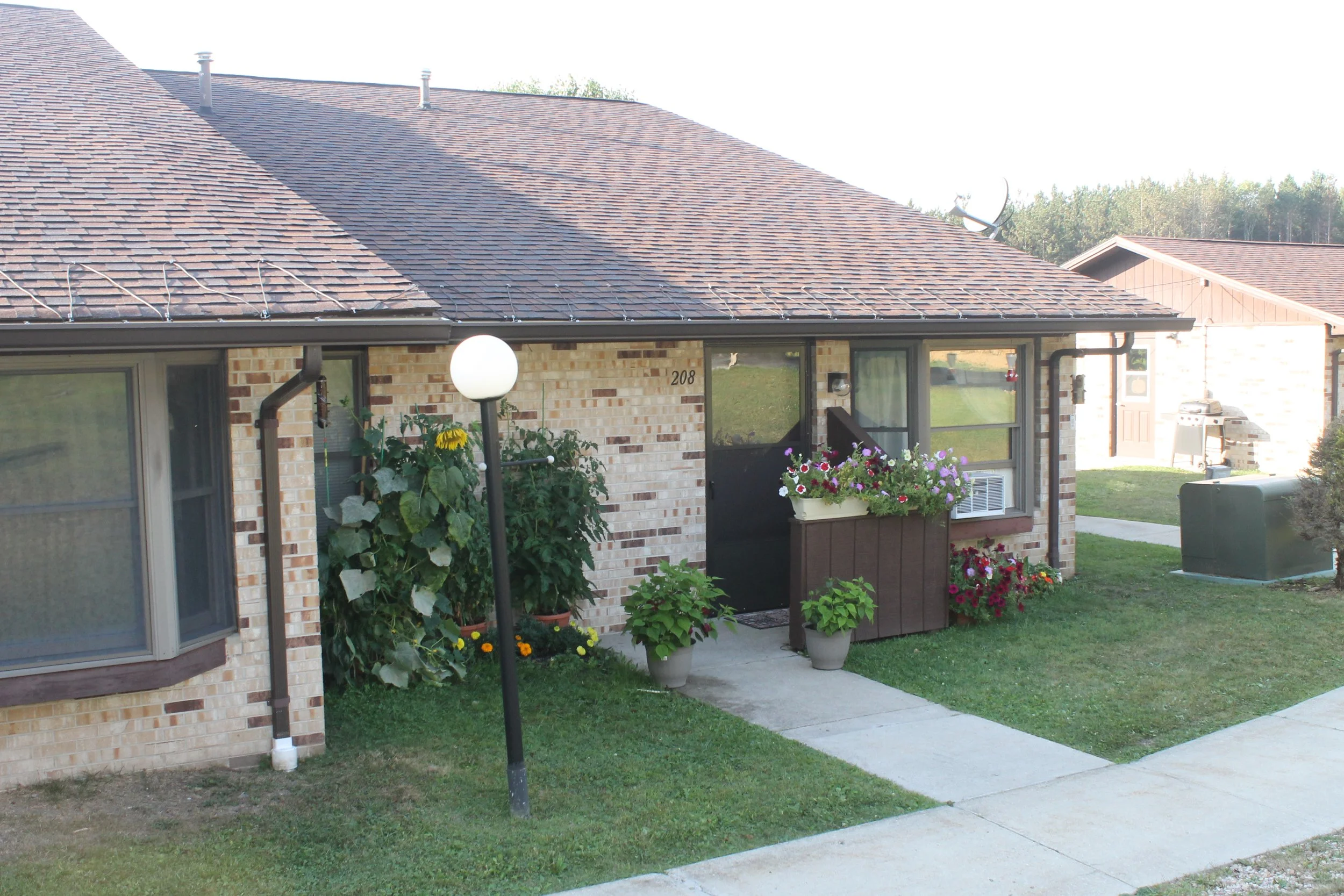 View of Pineview Apartments for low-income seniors and disabled residents. Front view of a brick house with a brown roof, potted plants near the entrance, a white outdoor lamp, and a sidewalk.