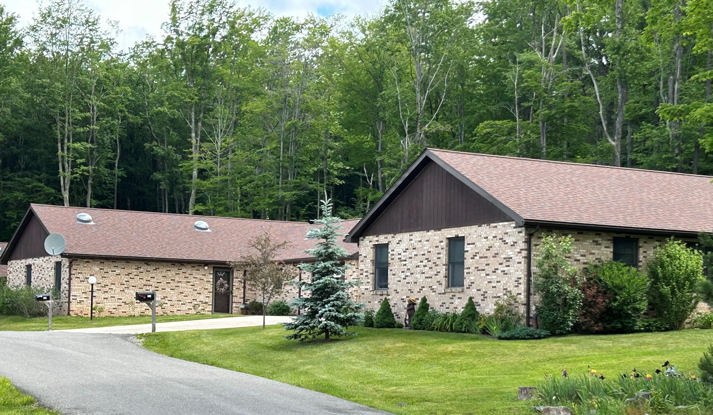 One of the villas at The Pines Community. Each villa has two units joined at the garage. A brick house with a gabled roof and a manicured lawn with trees and shrubs, set against a backdrop of dense green forest.