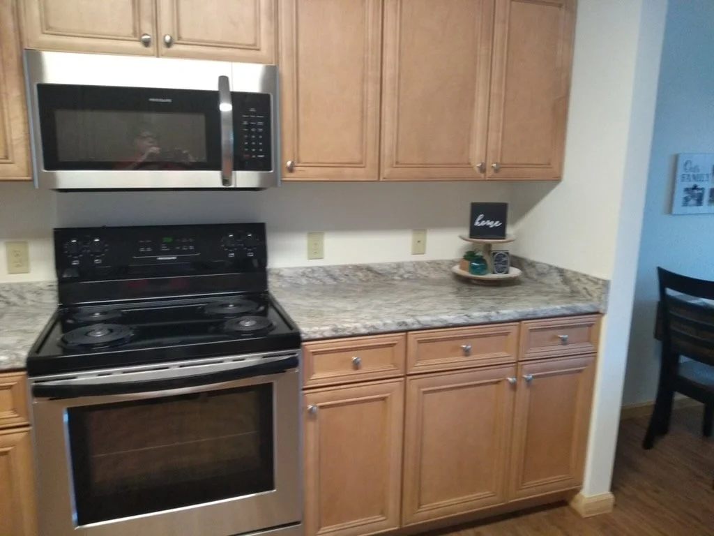 Kitchen with wooden cabinets, granite countertop, stainless steel microwave above a black stove, small decorative sign and plant on the counter, and part of a dining area with a black chair visible on the right.