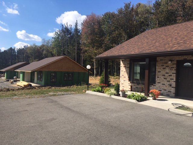 Our condos at The Pines. Four condos are inside each building. Residential area with a newly built house on the right with brick exterior and landscaping, a clear sky with some clouds, and a wooded backdrop.