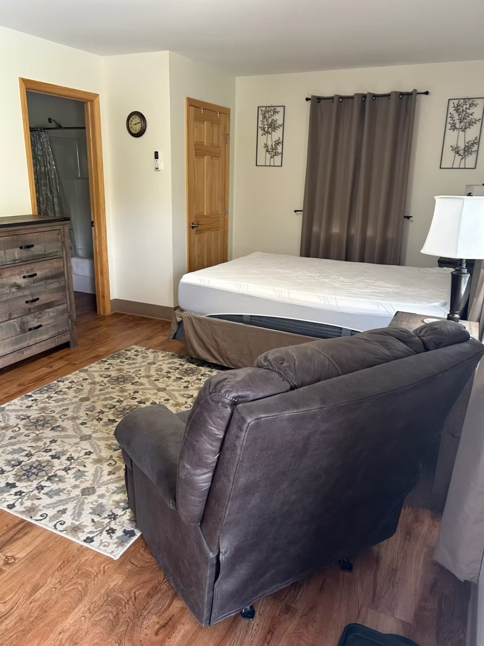 A studio apartment bedroom at the Rapids with a dark leather sofa, bed with white mattress, wooden dresser, closet with wooden door, gray curtains, and wall art featuring trees.