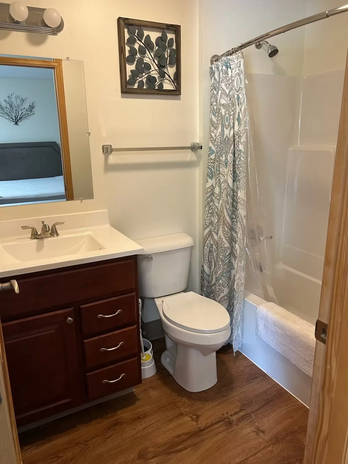 A Rapids Apartment bathroom with a wooden vanity, a mirror, a light fixture, a toilet, a shower with a patterned curtain, and framed artwork on the wall.