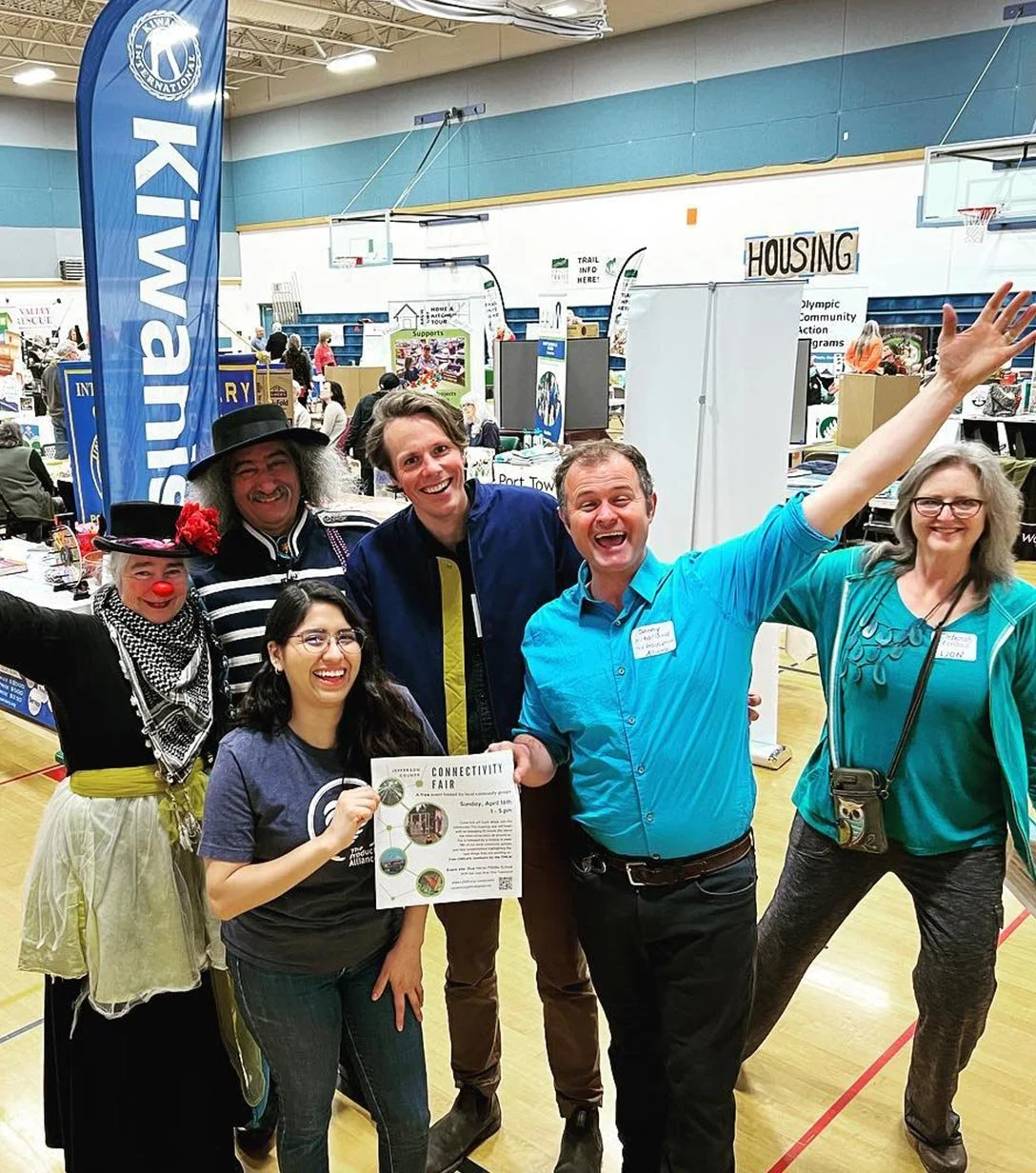 Group of six people posing cheerfully at a community event in a gymnasium with booths, banners, and signs in the background, including a large blue Kiwain banner.
