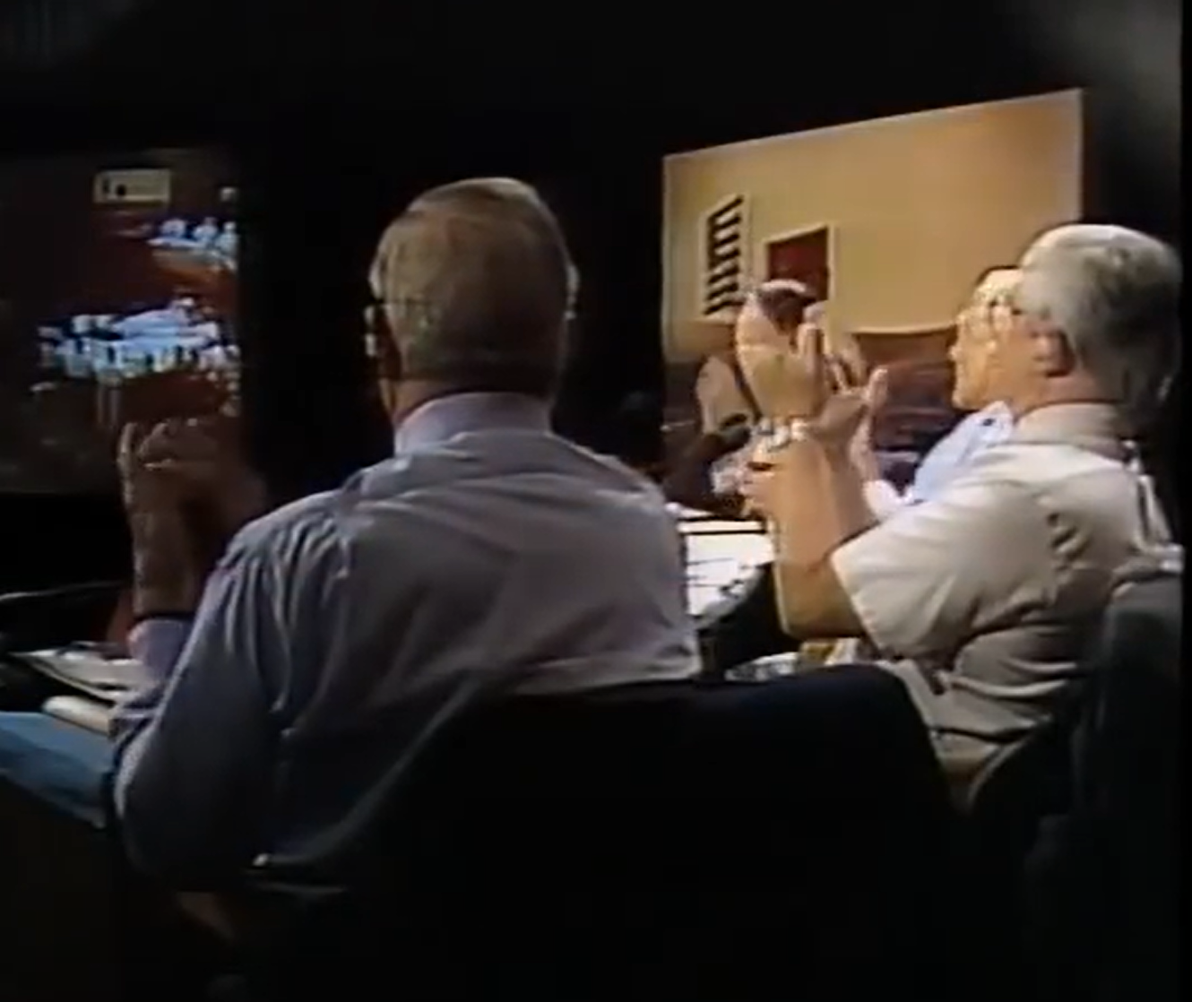 Three men sitting at a table in a dark room, watching a presentation or television screen showing a kitchen scene. They appear engaged and are gesturing with their hands.