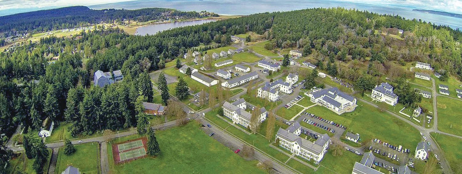 Aerial view of a small town or residential area surrounded by dense trees, with a river or lake in the background, showing streets, houses, parking lots, and a tennis court.