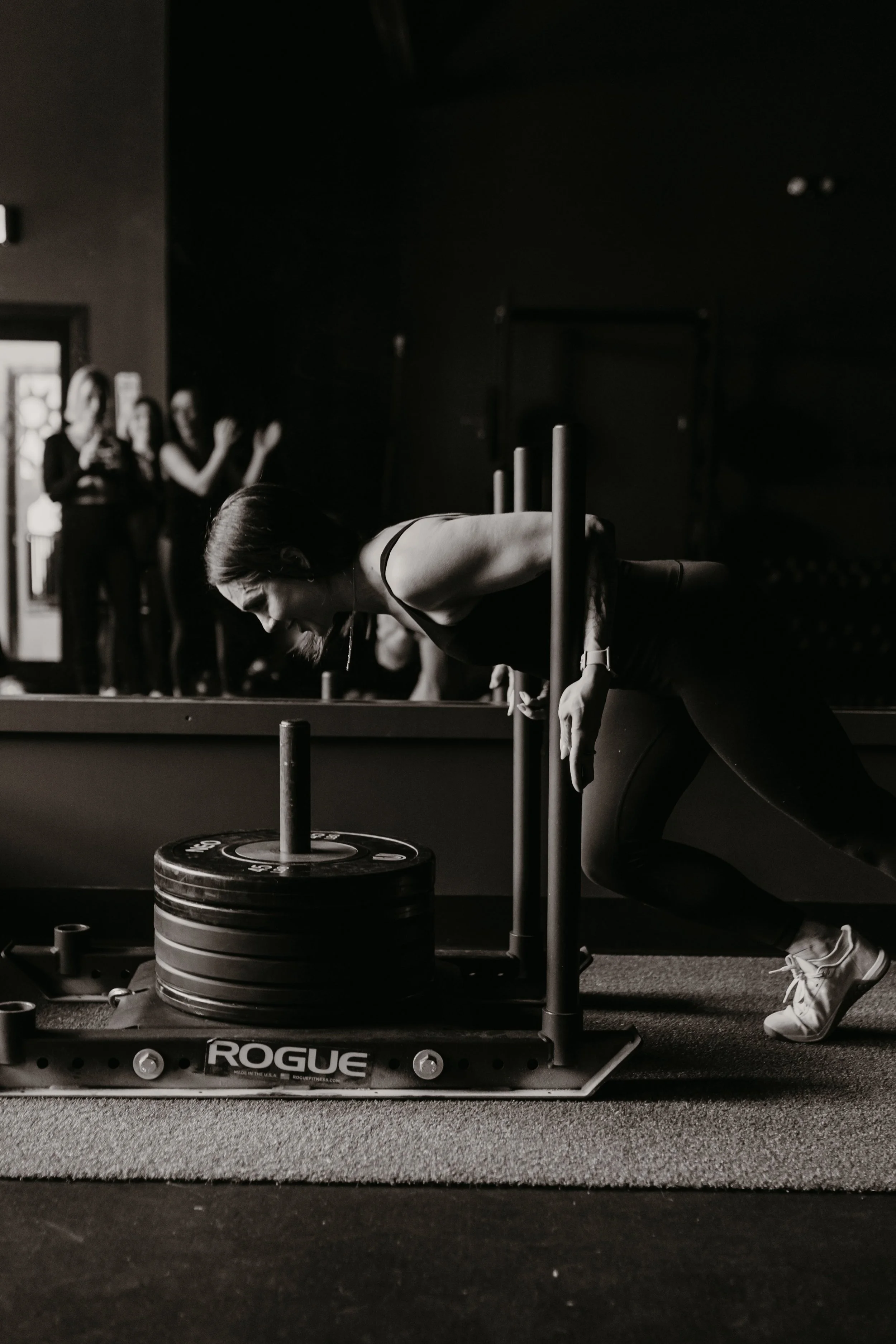 A woman in a gym performing a deadlift with weights, while three women in the background watch and clap.
