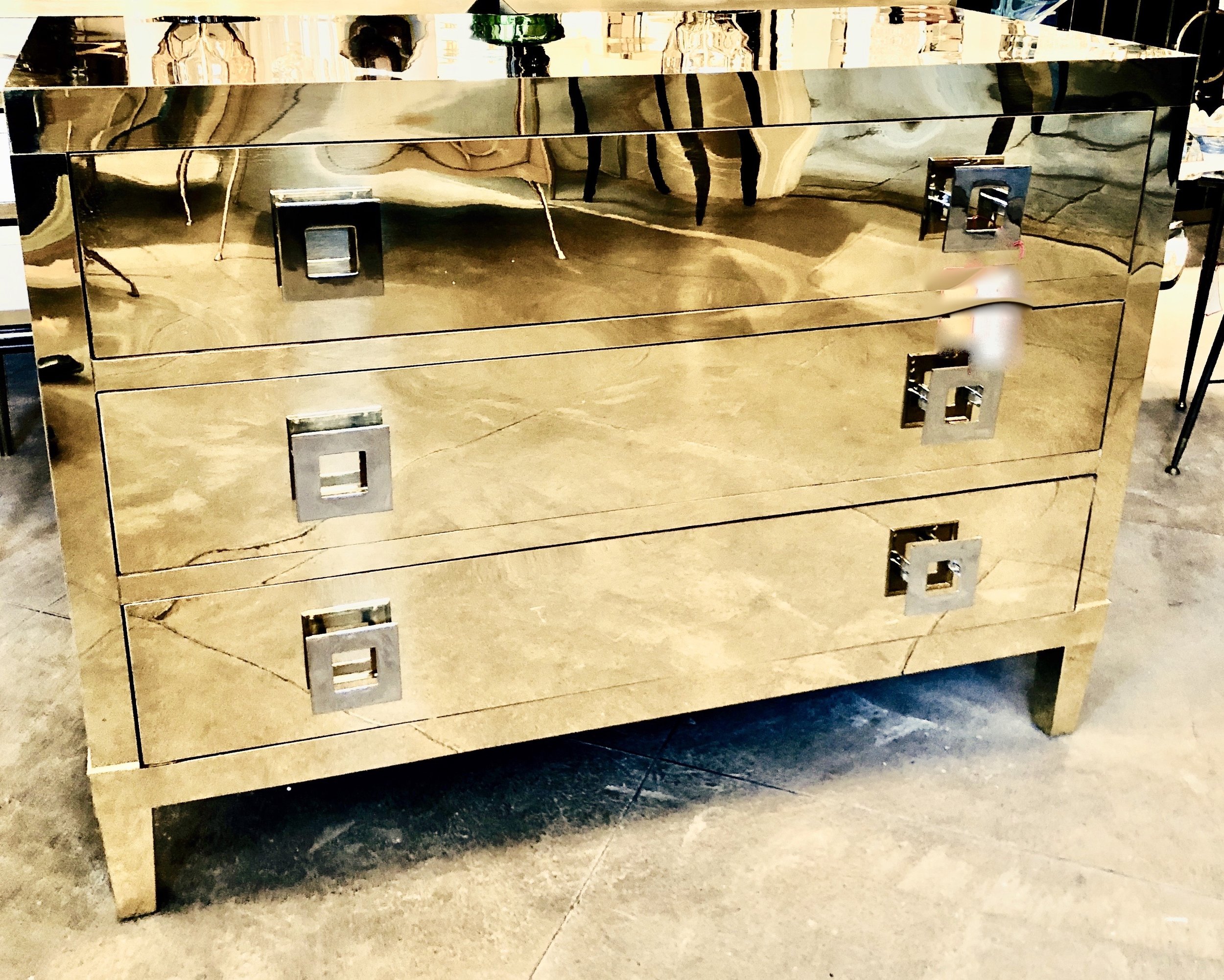 A mirrored dresser with four drawers and square handles, placed on a concrete floor in a store or warehouse setting.