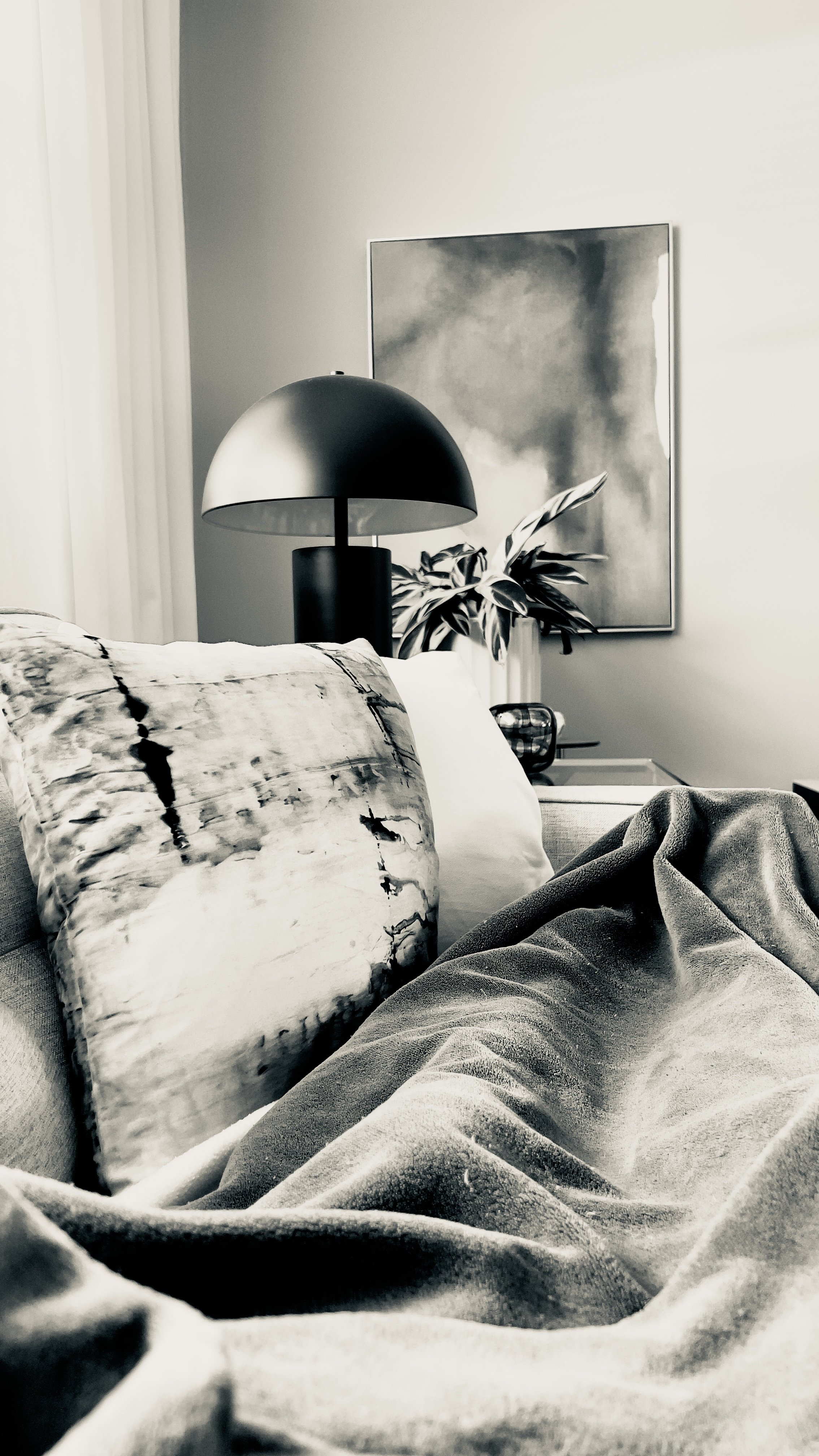 A living room scene with a grey linen sofa in the foreground, a black dome-shaped lamp, a potted plant, a piece of abstract art on the wall, and a side table.