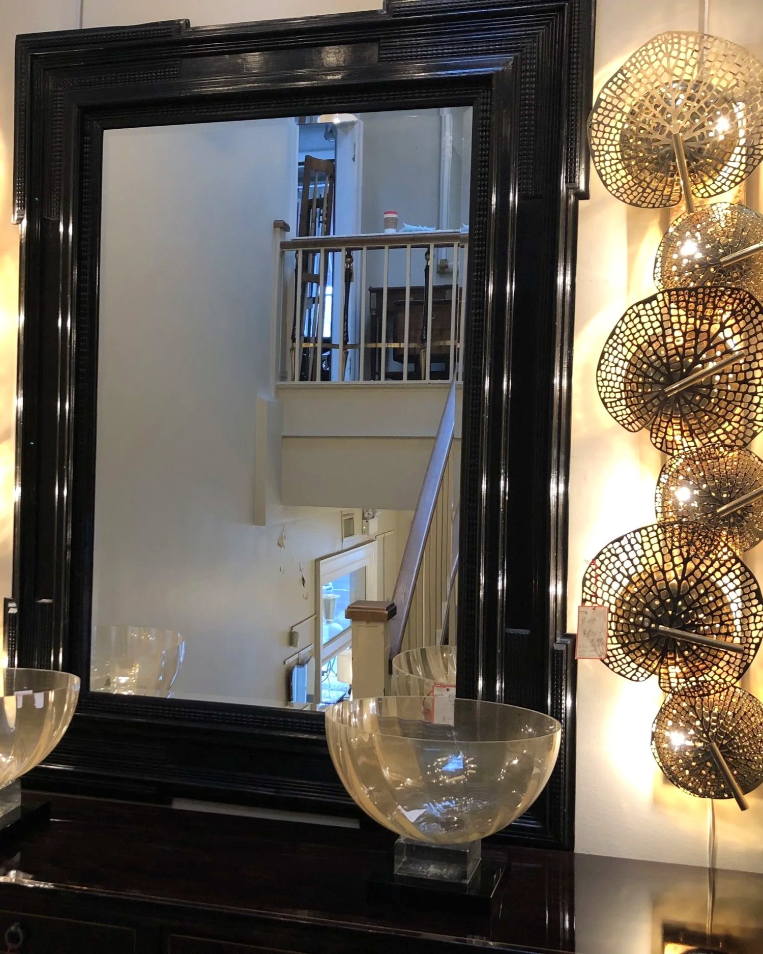 A large black-framed mirror reflects a stairway with a wooden railing and some furniture in the background. To the right of the mirror, there are four cylindrical, lattice-patterned wall lamps with warm lighting. In front of the mirror, there are three large, transparent glass bowls on a black surface.