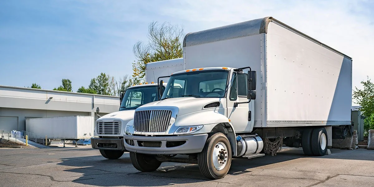 Two white box trucks parked outdoors on a paved surface with a building and trees in the background.