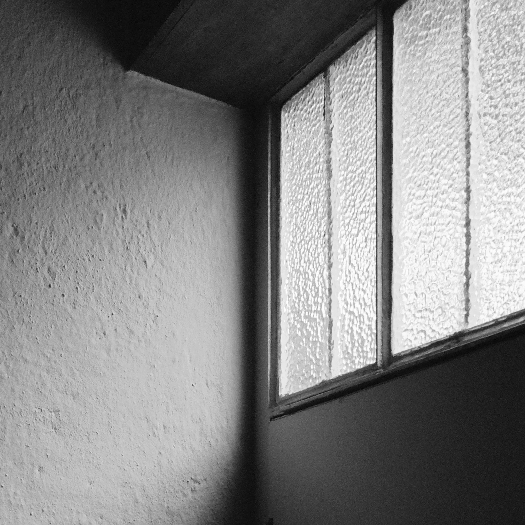 Black and white photo of a corner of a room showing a textured wall and a window with frosted glass panes.