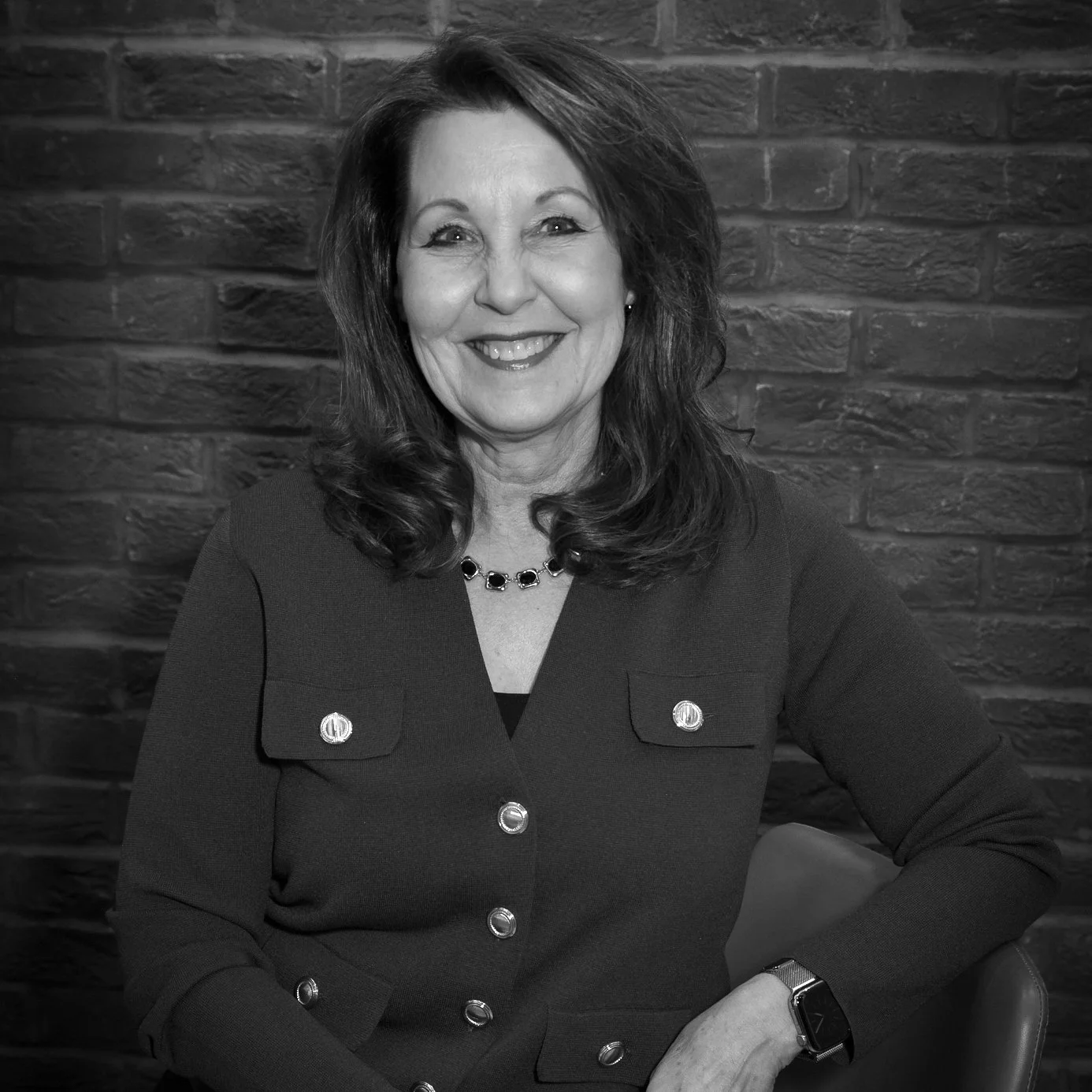 Black and white photo of a smiling woman with shoulder-length hair, wearing a dark blazer with large buttons, a necklace, and a watch, sitting against a brick wall background.